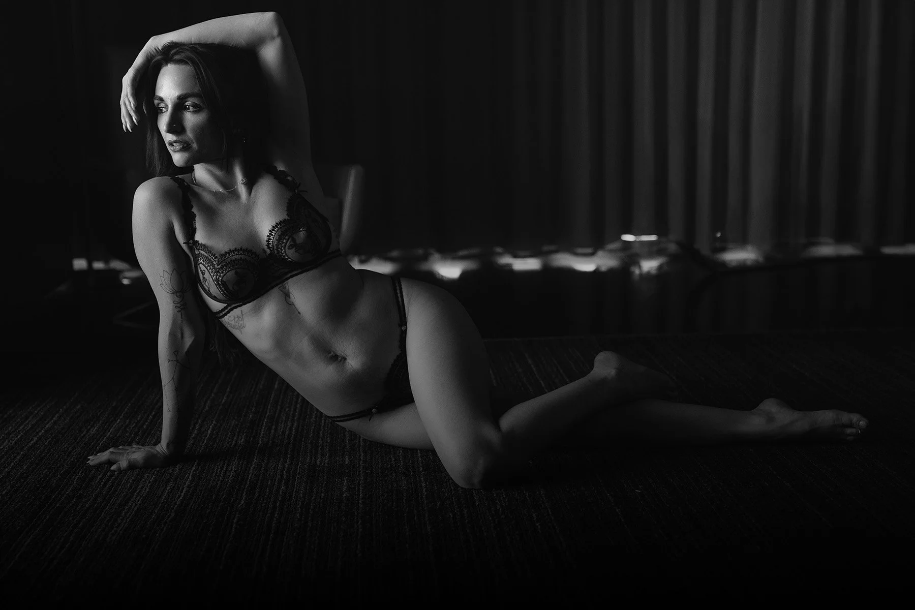 A woman in lingerie poses gracefully on a dark floor. The elegant black and white tone and soft lighting give the scene an artistic, dramatic feel.