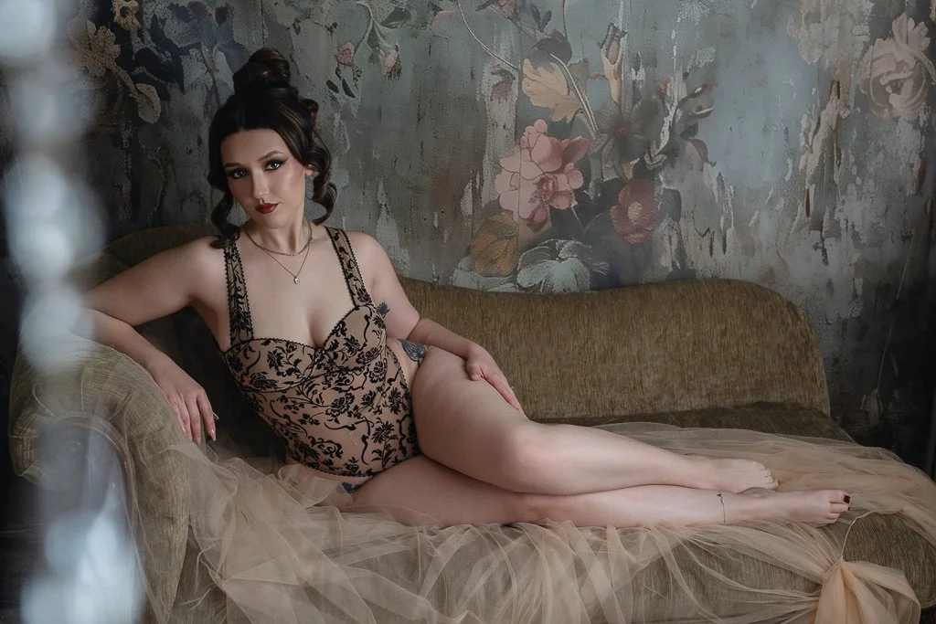 A woman in a vintage floral bodysuit reclines on a velvet chaise, surrounded by floral wallpaper and sheer fabric in a moody, elegant setting.