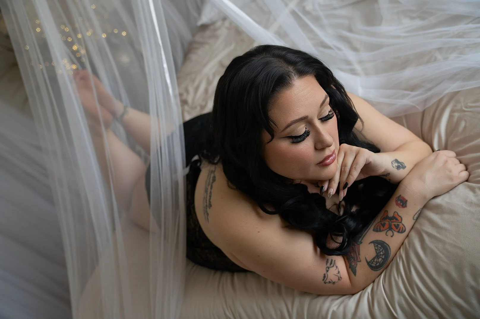 A woman with dark hair and tattoos lounges on a bed surrounded by sheer white fabric. She exudes a contemplative and serene vibe.