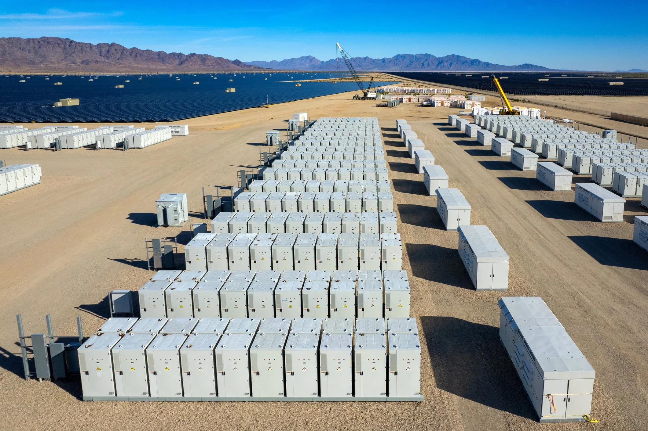 Solar panel farm with rows of energy storage units, some with cranes working, in a desert landscape with mountains in the background.