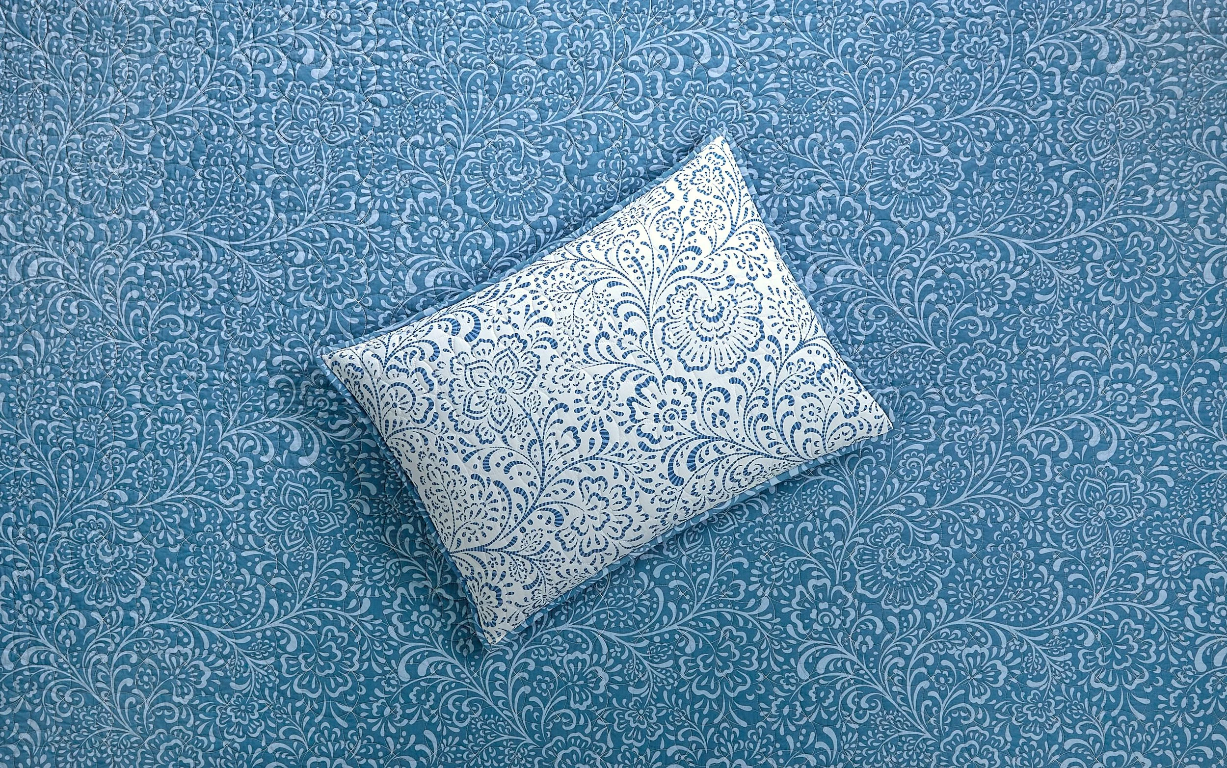 A white pillow with blue floral patterns on a blue floral bedspread.