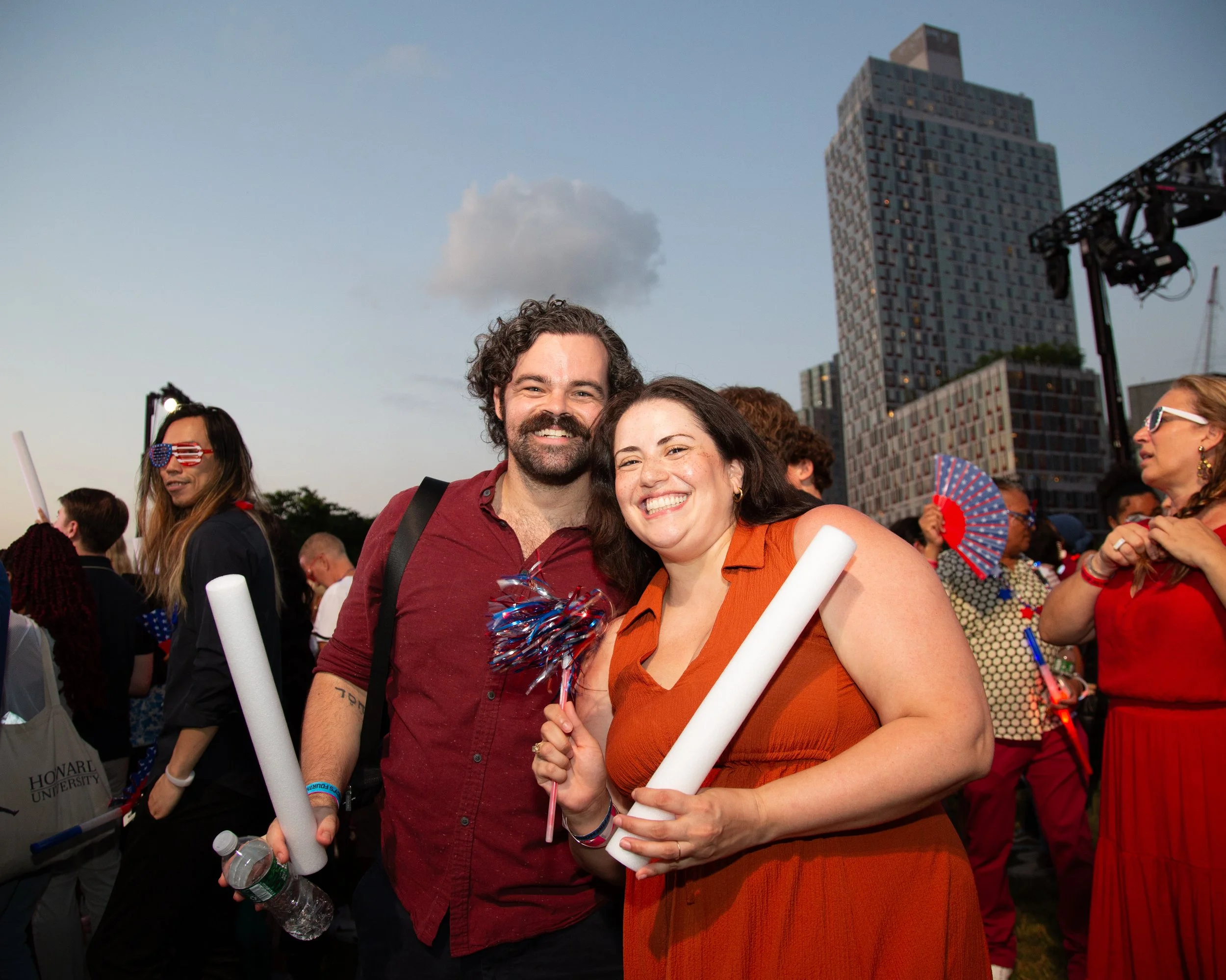 Smiling man and woman celebrating at an outdoor event, holding rolled-up papers, with other festive people in the background, city buildings, and a cloudy sky.