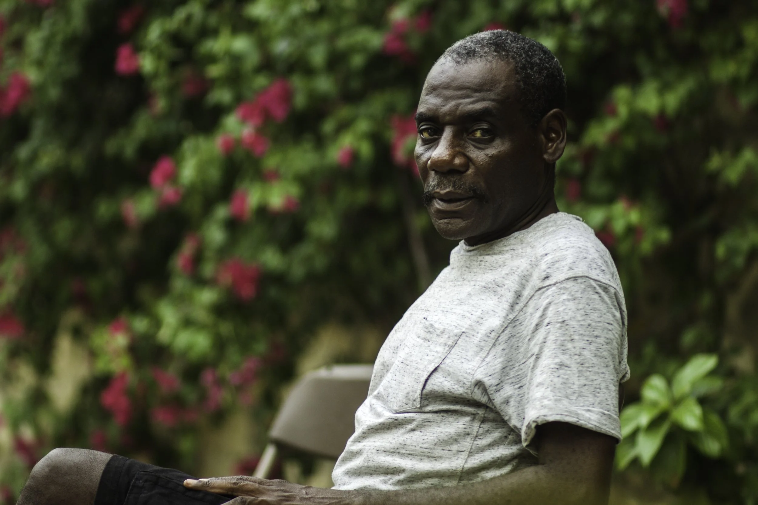 A middle-aged man sitting on a side view chair outdoors, wearing a light gray T-shirt and black shorts, with a background of green foliage and pink flowers.