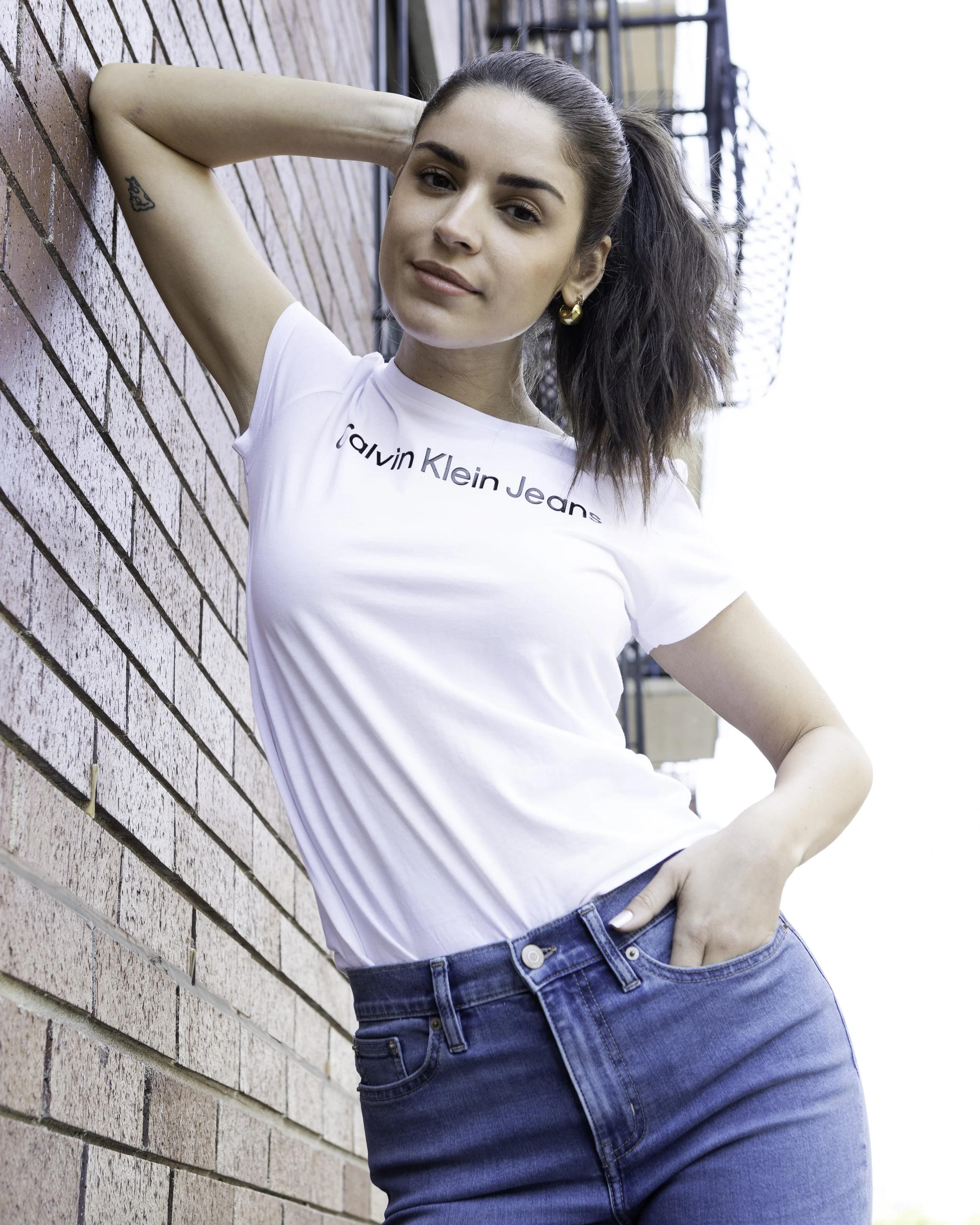Young woman with brown hair tied in a ponytail posing against a brick wall, wearing a white Calvin Klein Jeans t-shirt and blue jeans. Macy's Lifestyle 