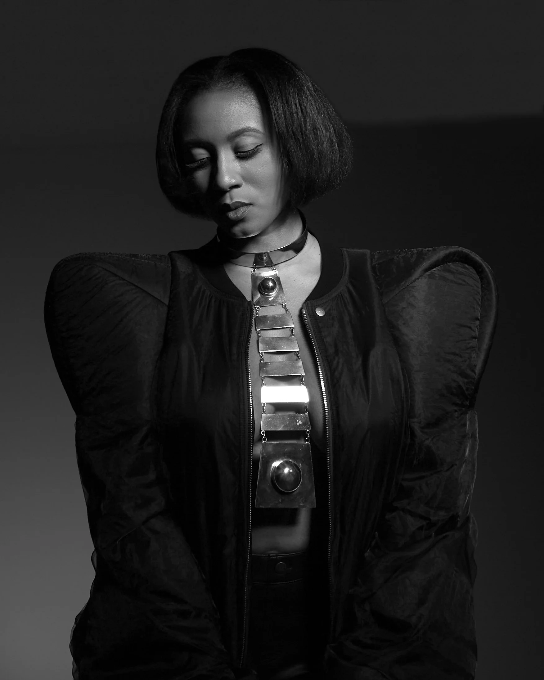Black and white photo of a woman with short hair, wearing a bomber jacket and metallic jewelry, looking down with a contemplative expression.
