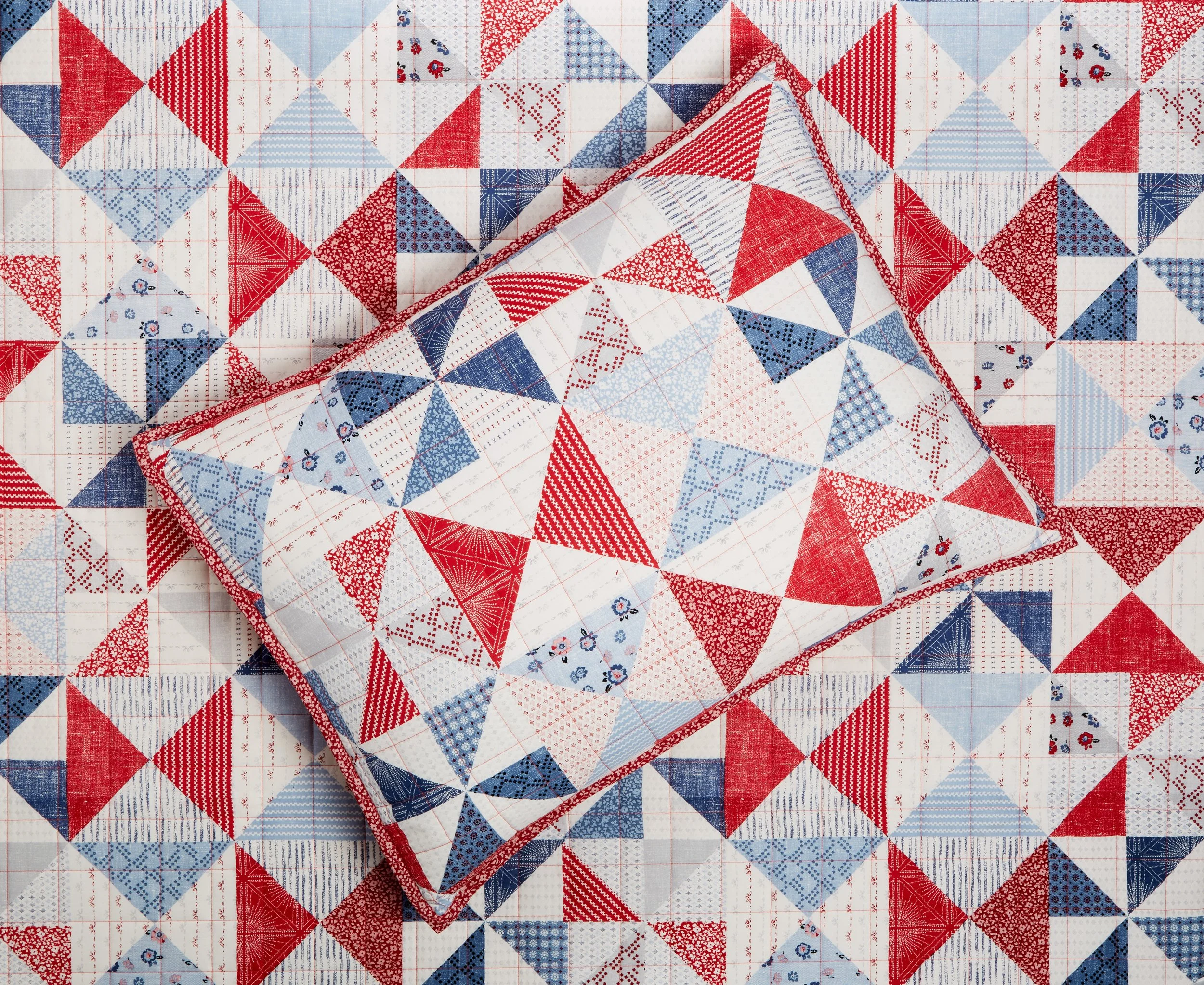 A quilted fabric with a patchwork pattern of red, blue, and white triangles and squares. A pillow resting on the quilt matches the pattern.