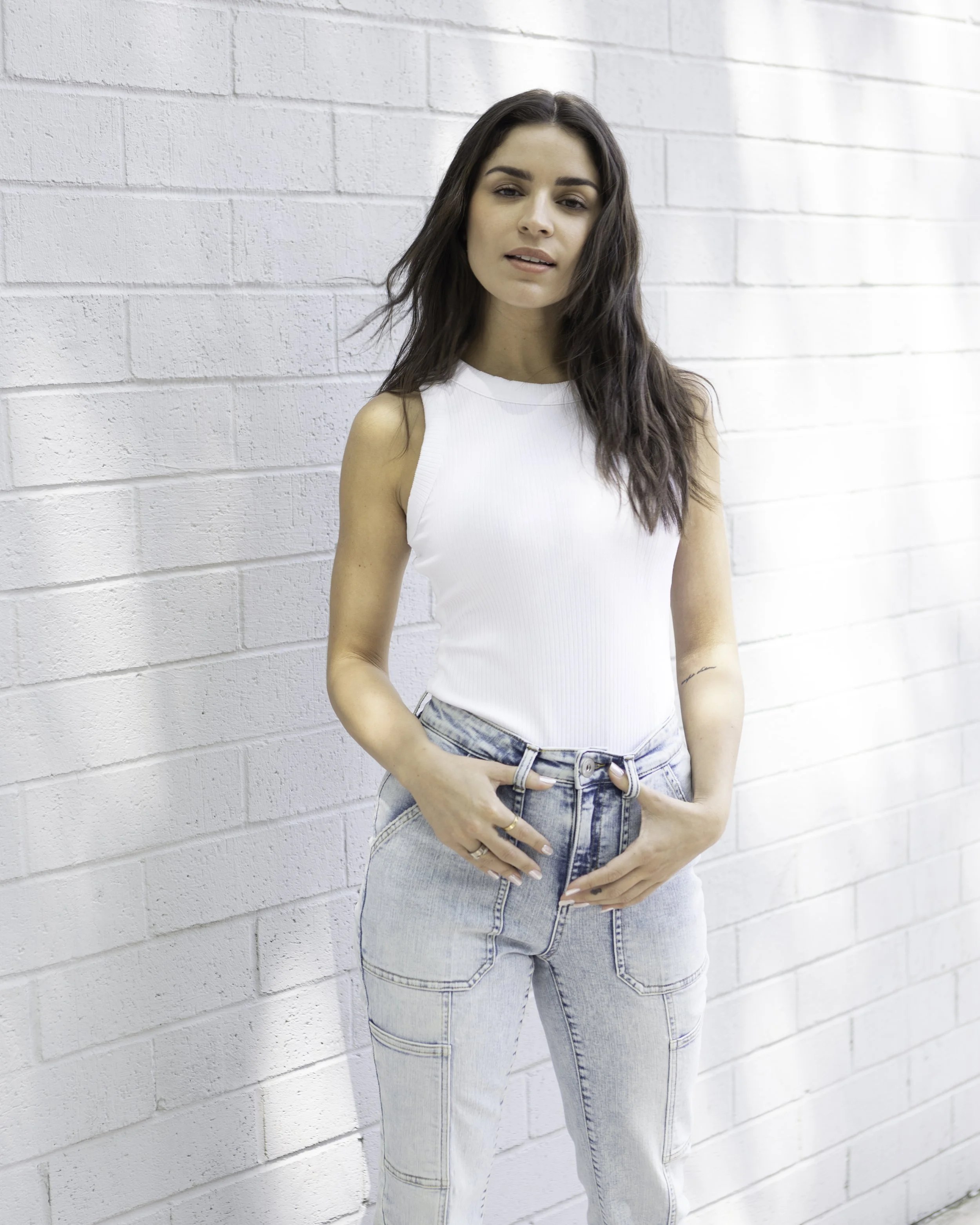 Young woman with wavy brown hair wearing a sleeveless white top and light-wash jeans, standing against a white brick wall. Macy's Lifestyle 