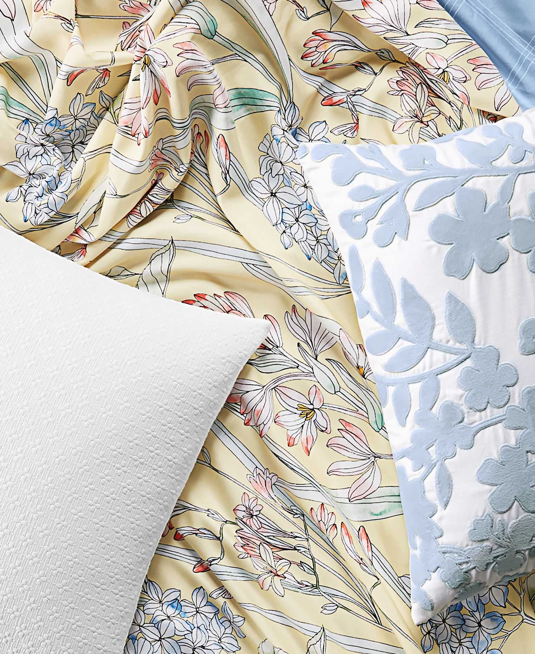 Close-up of a bed with floral patterned yellow duvet, a white pillow with textured pattern, and a pillow with light blue embroidered floral design.