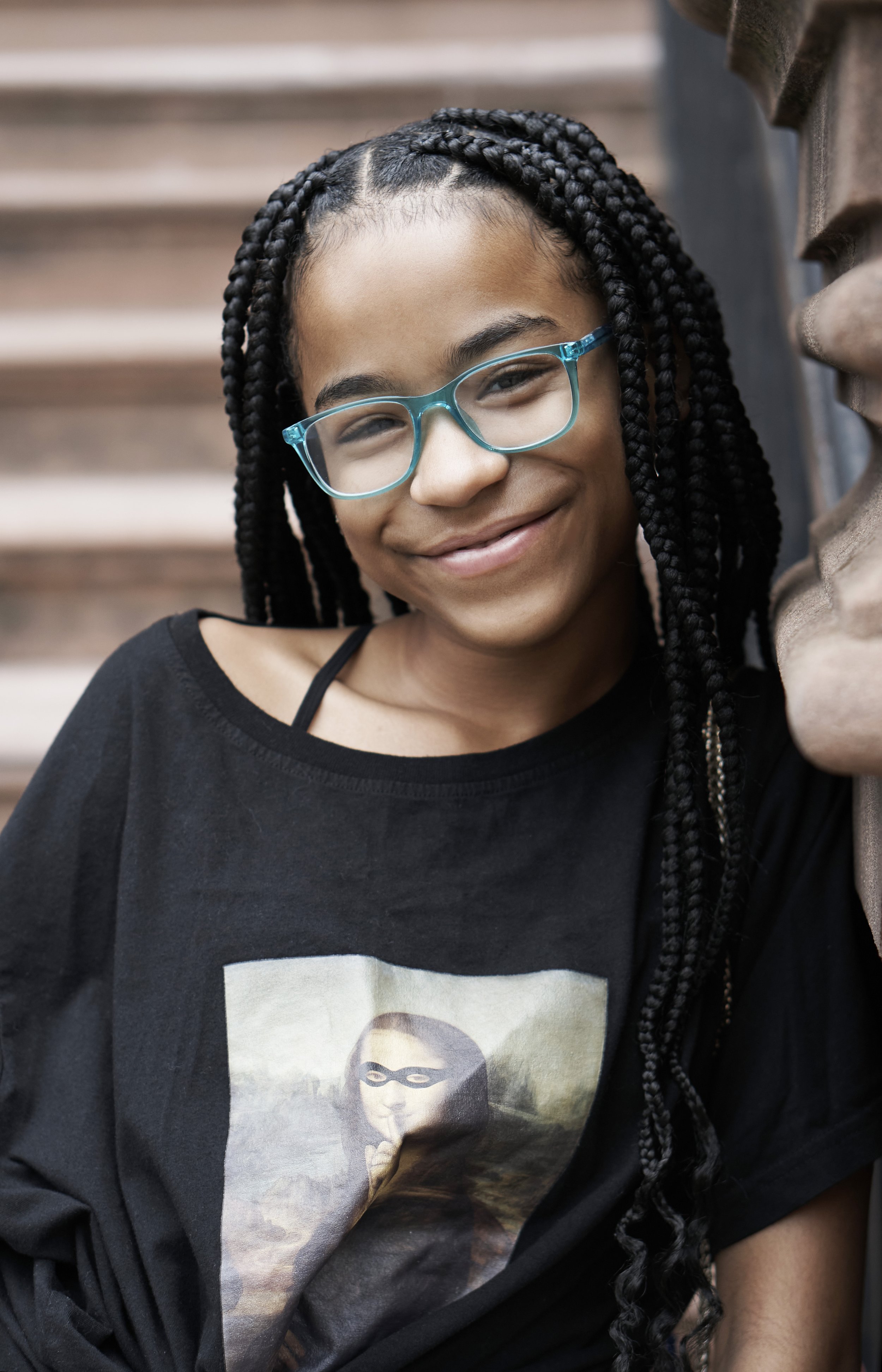 A young woman with braids and glasses smiling outdoors, leaning against a brick wall, wearing a black T-shirt with a Mona Lisa print. Harlem 