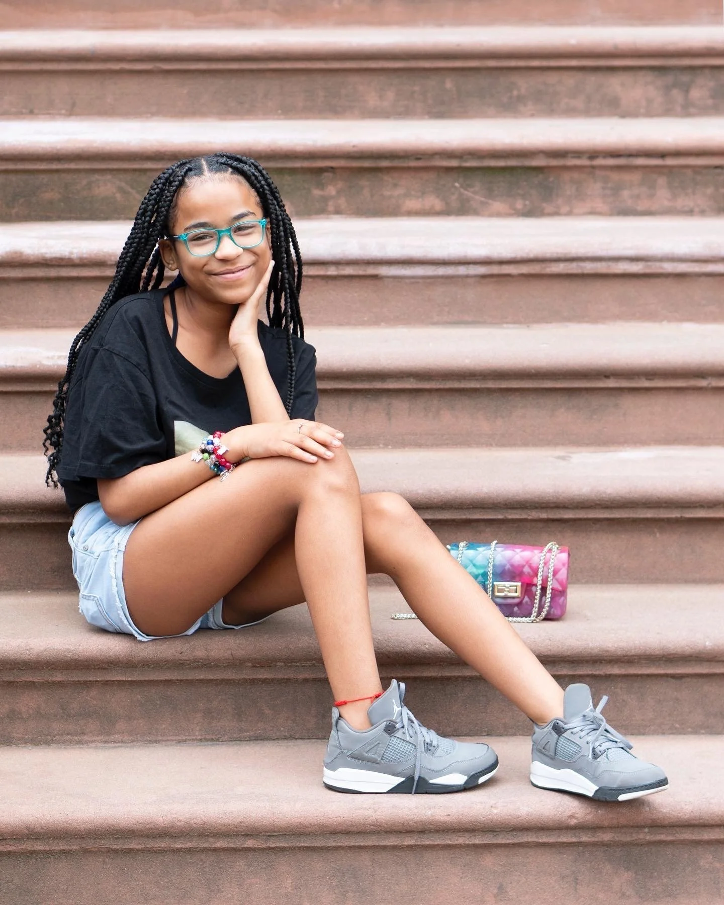 A young girl with glasses, braided hair, and colorful jewelry sitting on outdoor steps, smiling with her hand resting on her cheek. She has a backpack and is wearing casual clothes and sneakers. Harlem 