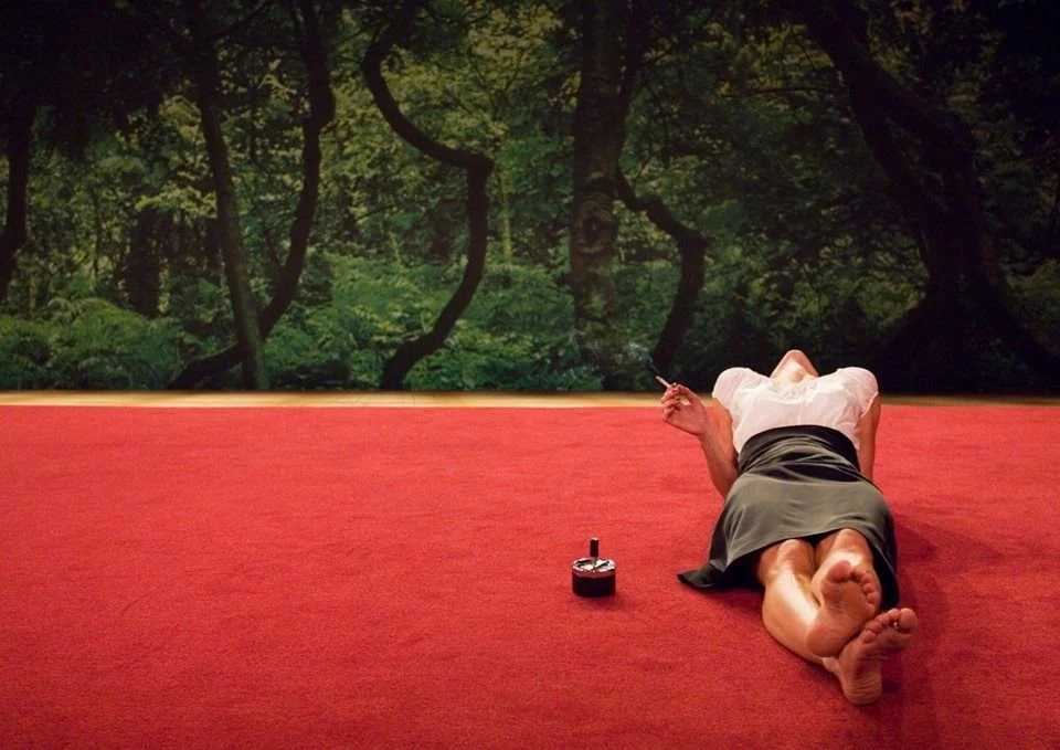 Person lying on red carpet in front of a forest background, holding a cigarette and an ashtray nearby.