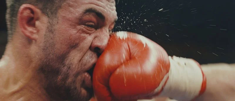 A boxer punches with a clenched red glove, his face close to the glove, sweat splashing.