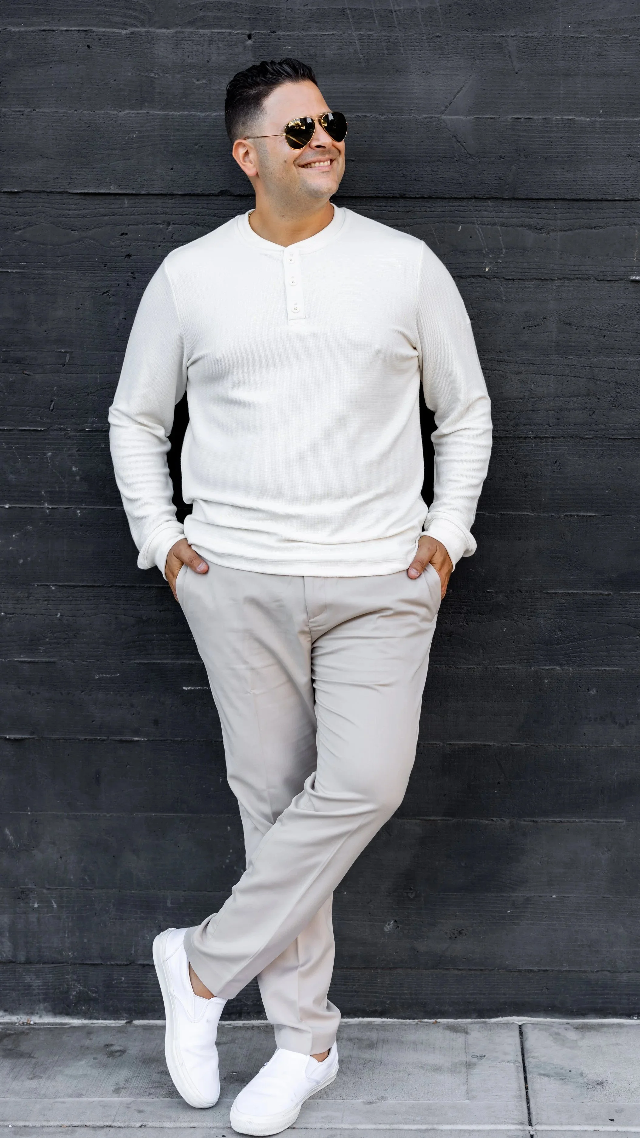 A man wearing sunglasses, a white long sleeve shirt, beige pants, and white sneakers leaning against a black wooden wall, smiling and with hands in pockets.