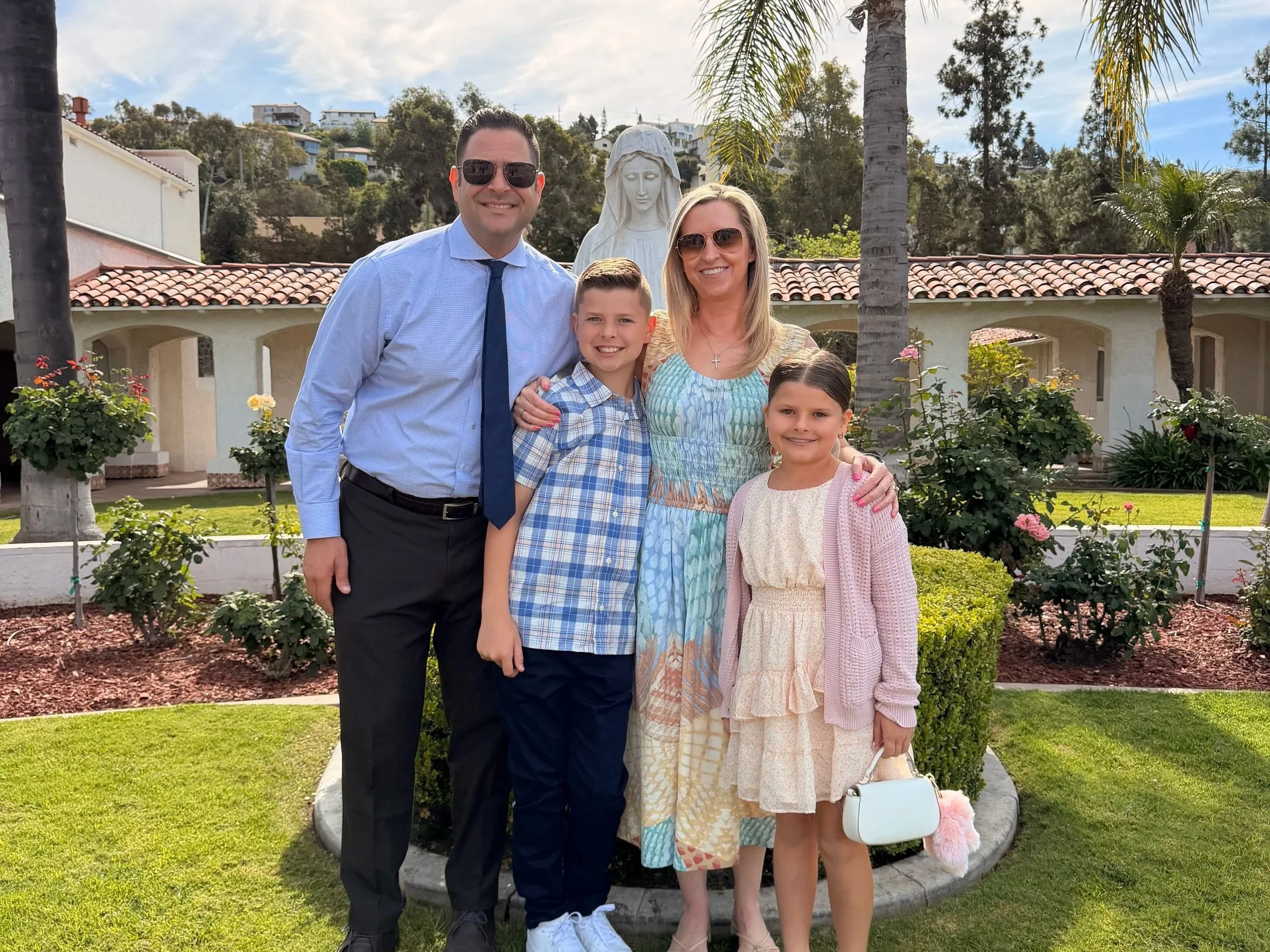 A family of five standing together outdoors near a statue of Mary. The family consists of a man, woman, two girls, and a boy, all smiling and dressed casually. The background features trees, flowering bushes, and a building with a red-tiled roof.
