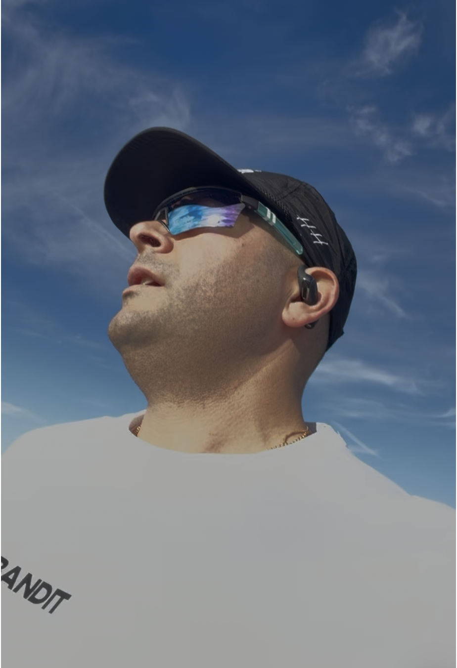 Man wearing a black cap, reflective sunglasses, and wireless earbuds, looking upward against a blue sky with clouds.