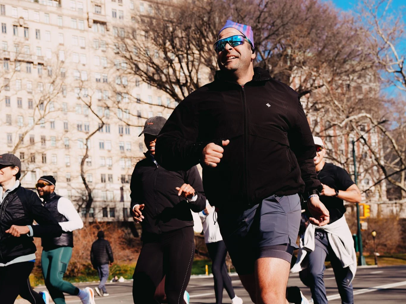 Group of people jogging outdoors in an urban park during the day, with a man in front wearing sunglasses and a colorful headband, trees, and a high-rise building in the background.