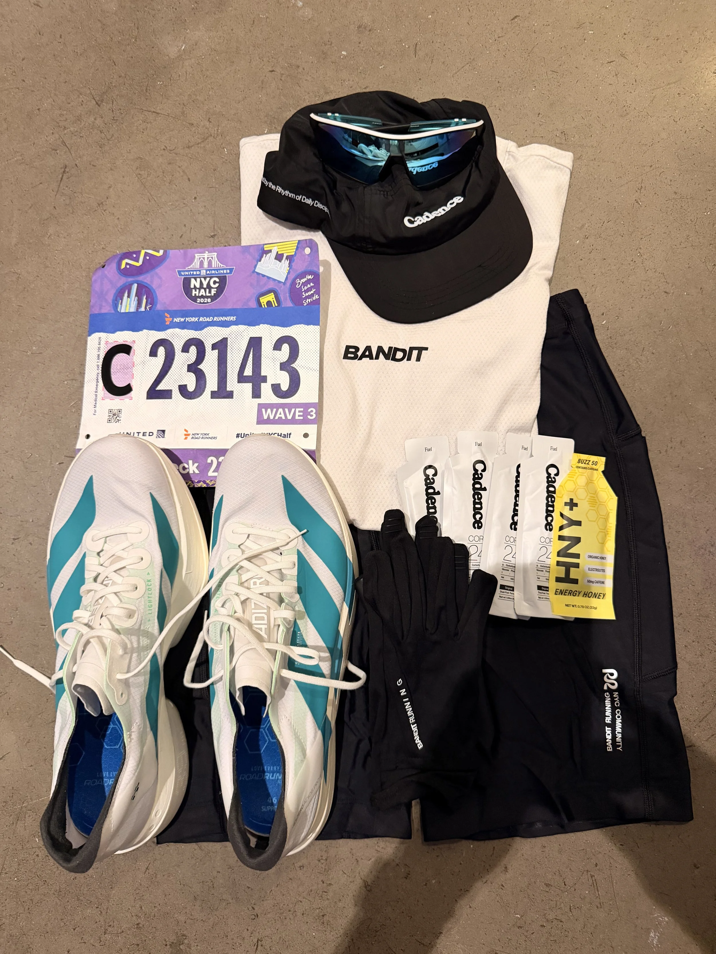 Collection of running gear including a pair of white and turquoise running shoes, a race bib number 23143, black gloves, a black cap with sunglasses on top, a white towel with a logo, several energy gel packets, and a black running bag, all arranged on a concrete floor.