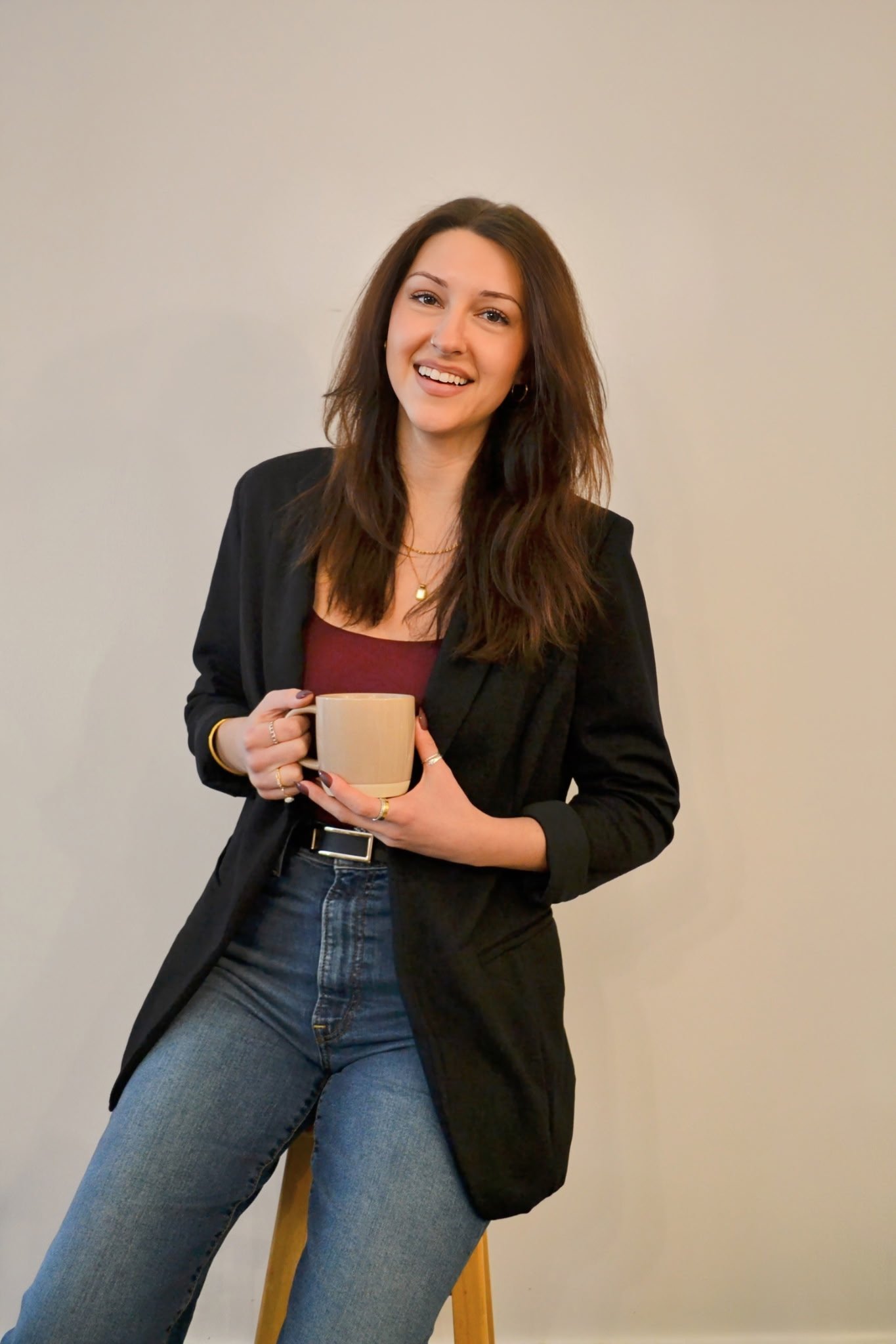 A young woman with brown hair smiling, sitting on a wooden stool, holding a beige coffee mug, wearing a black blazer, maroon top, and blue jeans against a plain light background.