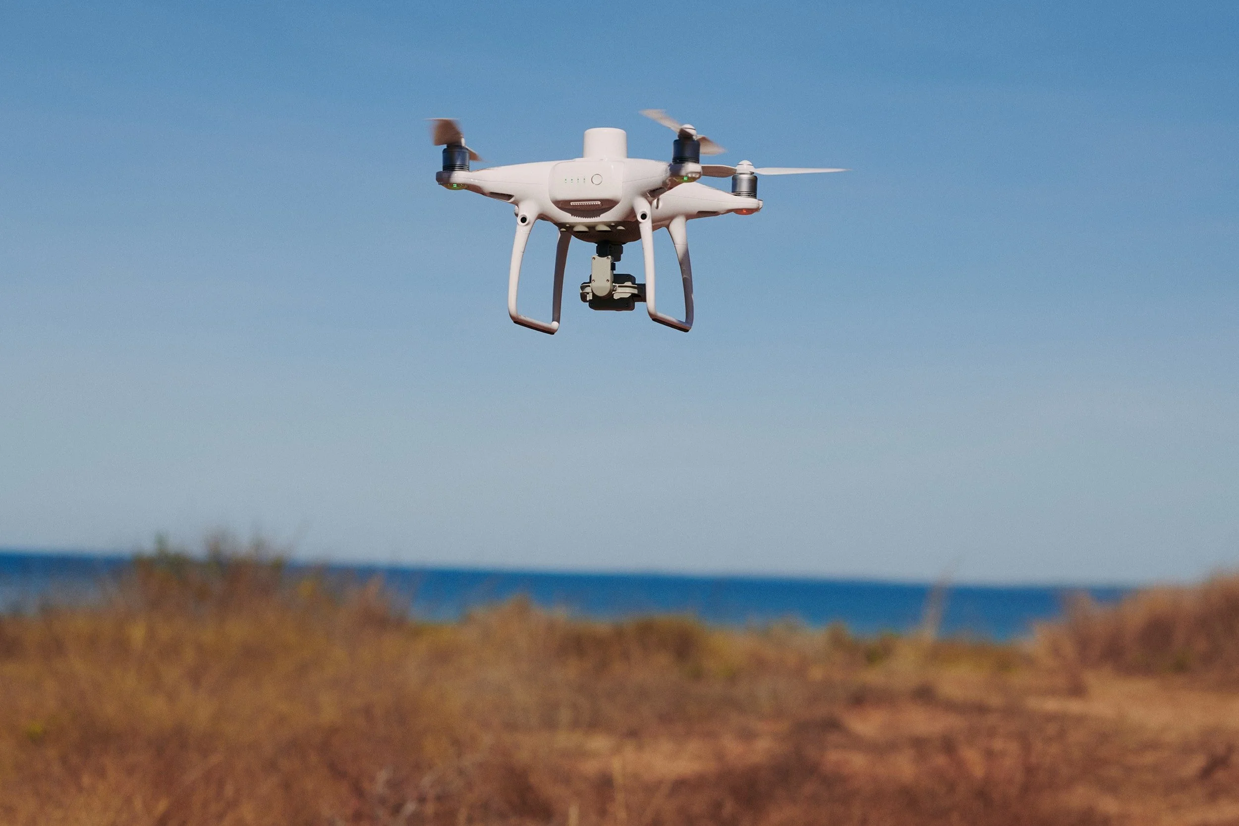 A white drone flying over a grassy area near the coast with blue sky in the background.