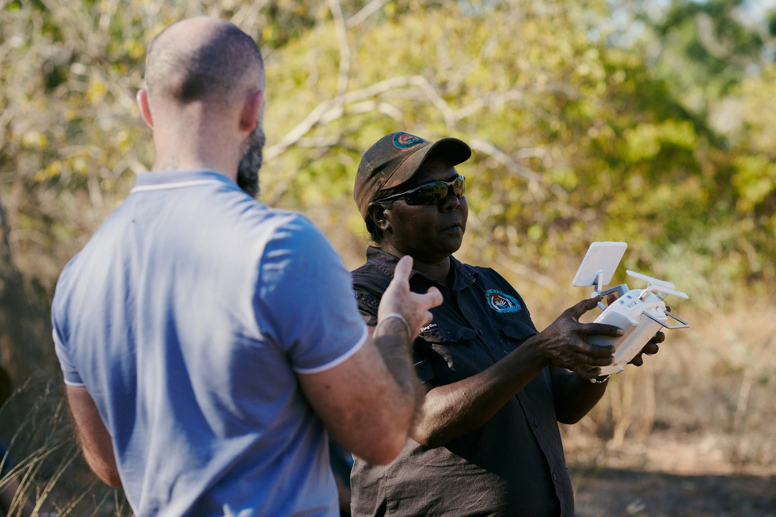 Two people outdoors using a drone controller. They are surrounded by trees and greenery.