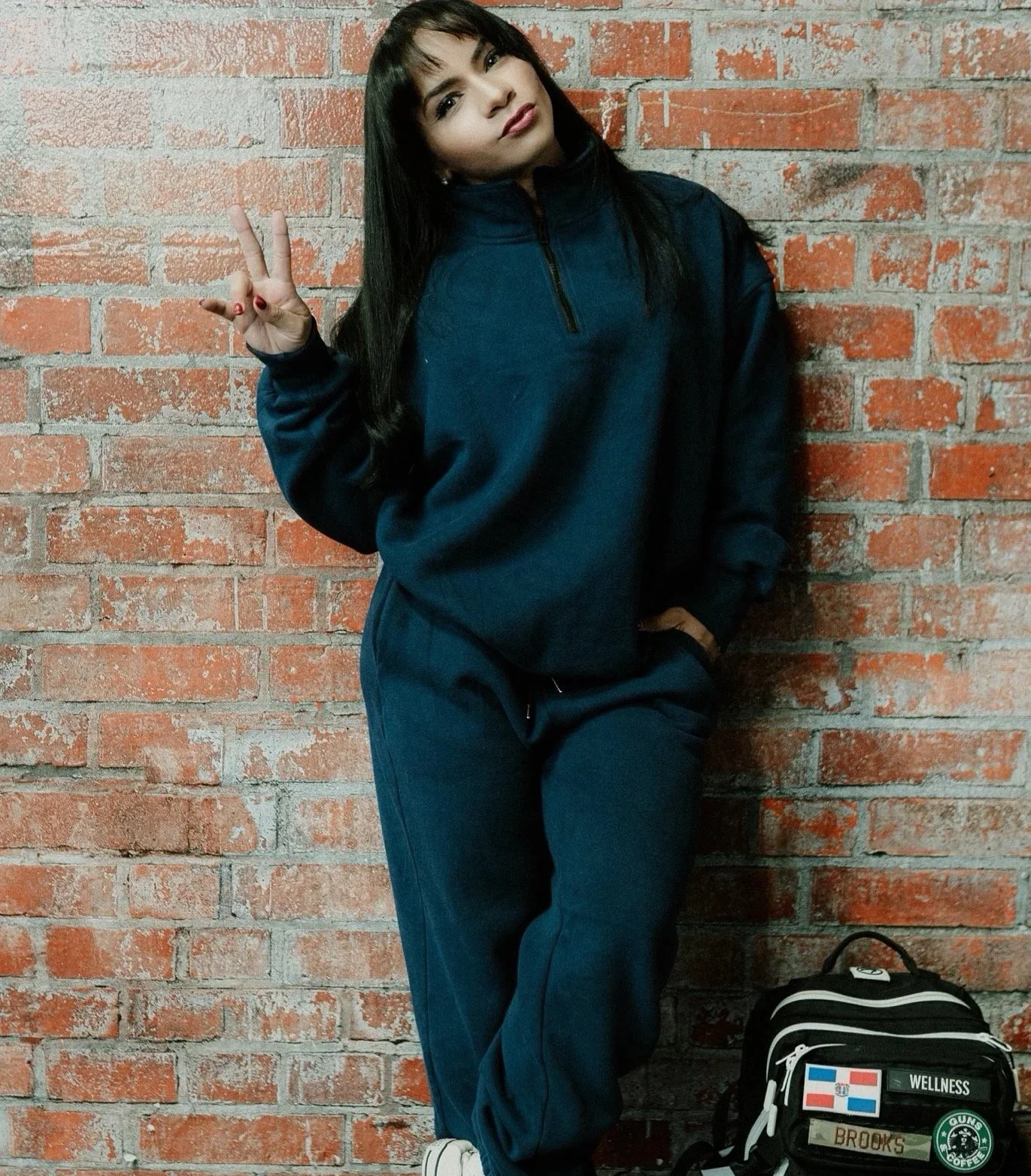 Young woman with dark hair and bangs wearing a navy blue tracksuit, standing against a brick wall, making a peace sign with her right hand, and standing next to a black backpack.