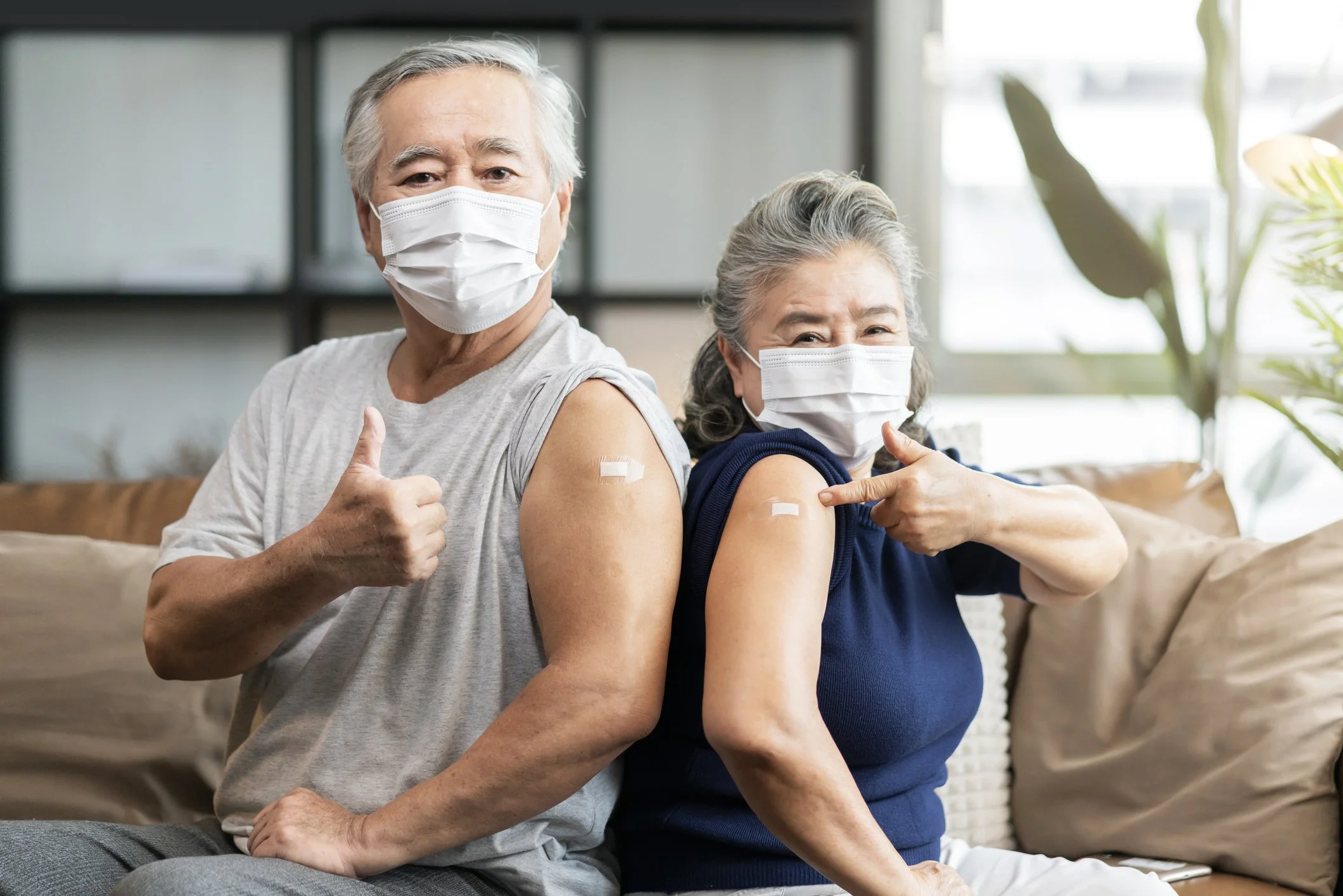Two older adults wearing face masks showing bandages on their upper arms, sitting back-to-back on a couch, indicating they have been vaccinated.