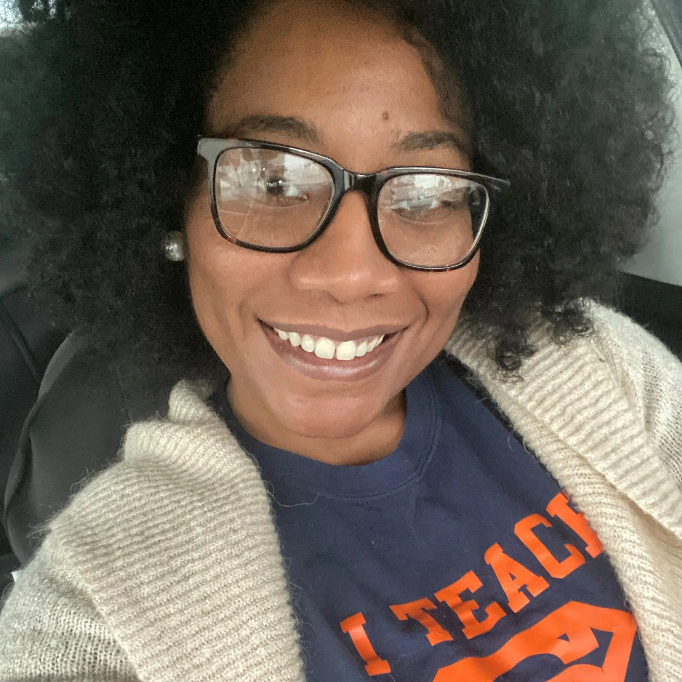 A woman with glasses smiling, wearing a gray sweater and a blue shirt with orange text, sitting in a vehicle.