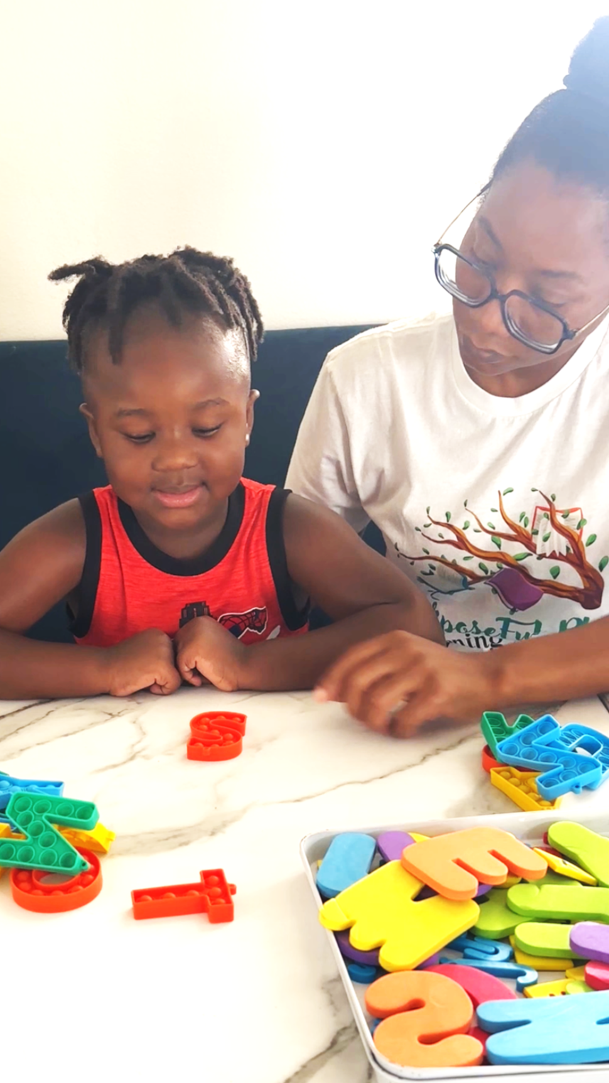 A young child and an adult woman sitting at a white marble table, playing with colorful foam alphabet and number puzzles. The child is wearing a red sleeveless shirt and the woman is wearing glasses and a white T-shirt with a tree and bird design.