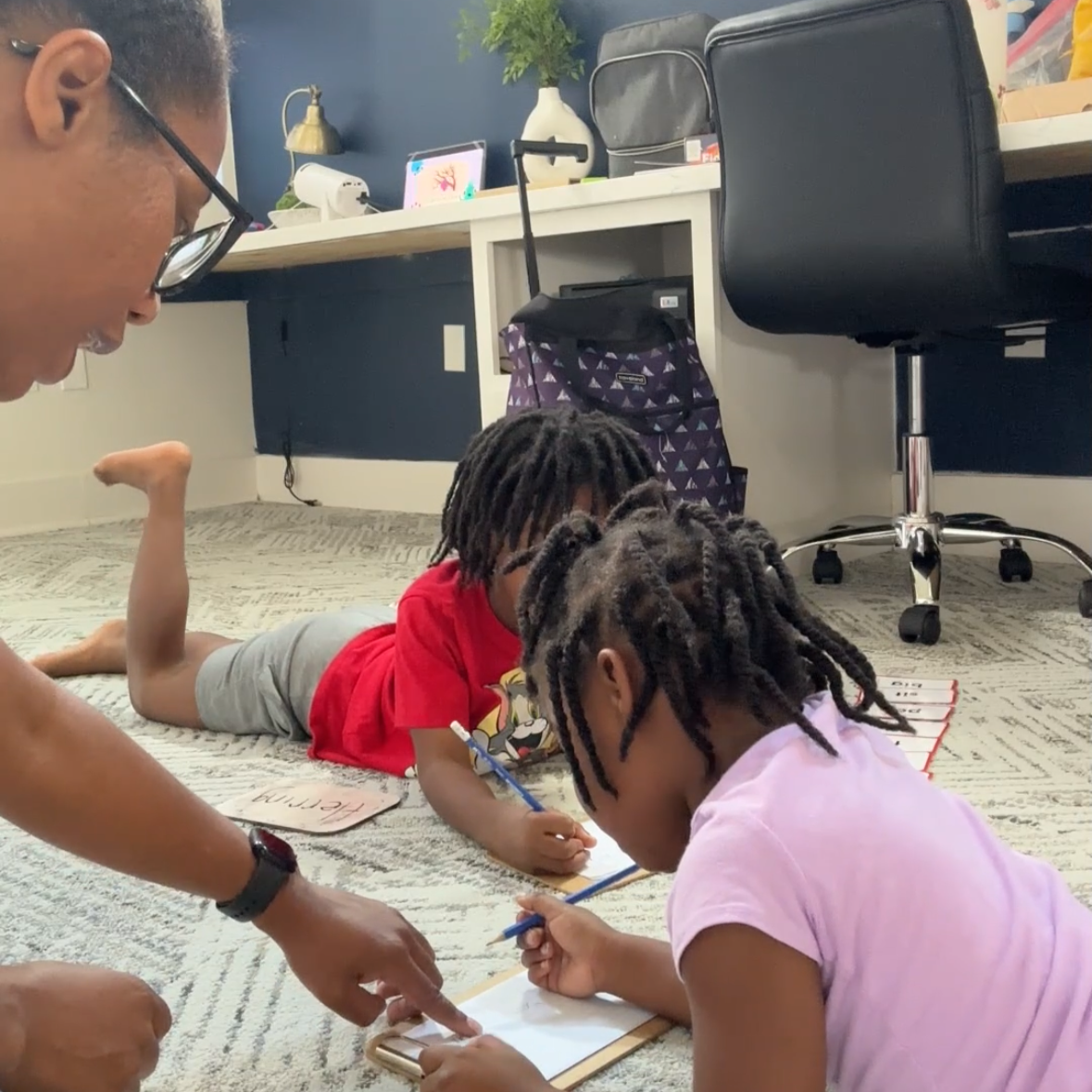 Two young children lying on the carpeted floor writing on clipboards with a teacher pointing at one of the clipboards. There are backpacks, a desk, and an office chair in the background.