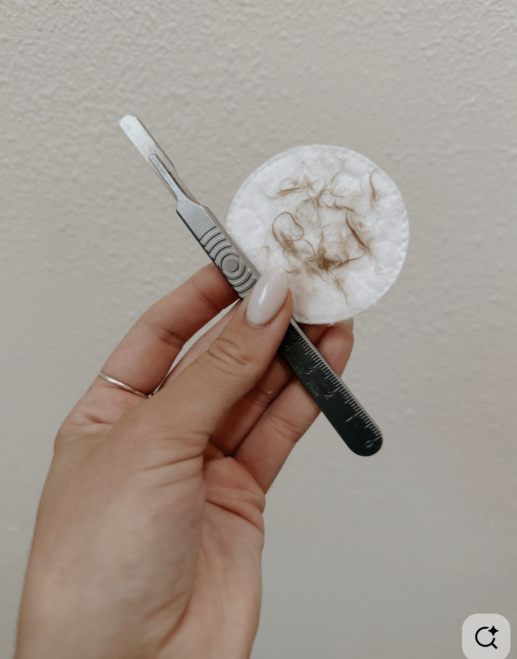 A hand holding a small metal tool and a round cotton pad on a neutral background.