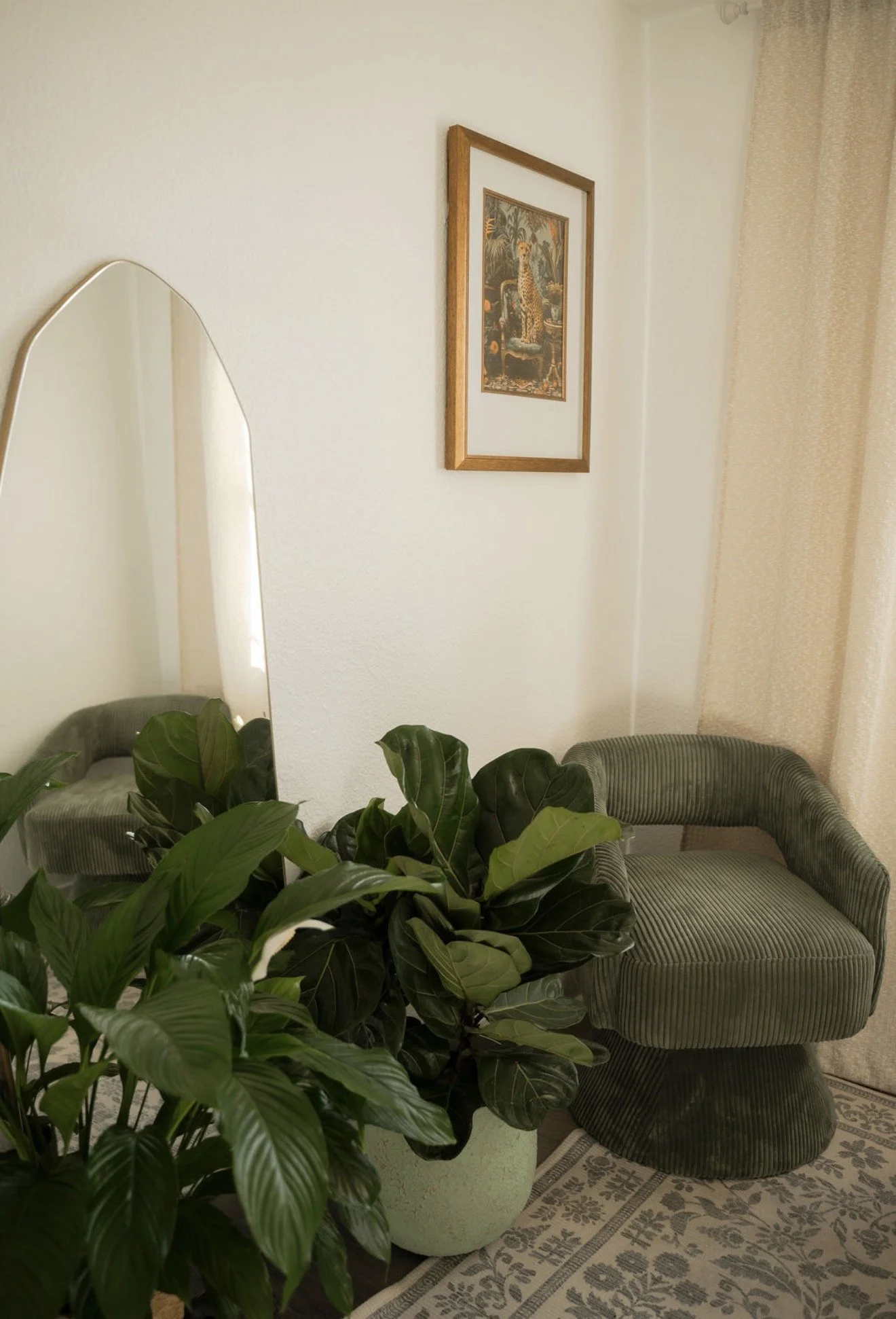 A room corner with a tall lush green potted plant, a green velvet armchair, a large oval mirror, a framed wall art of a jungle scene with a leopard, and beige curtains.