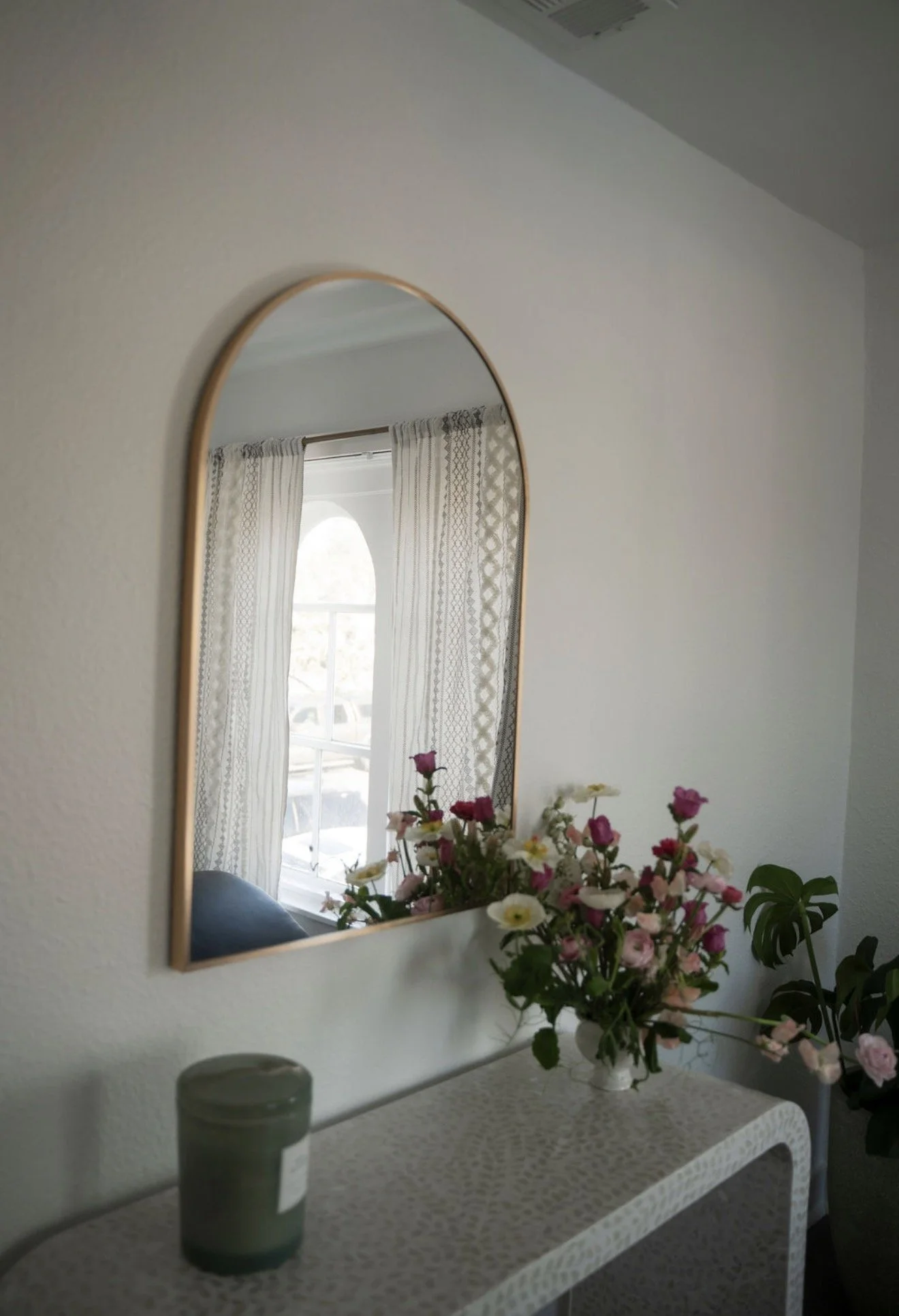 Interior view of a room showing a mirror, a window with patterned curtains, a bouquet of pink and white flowers on a table, and a green potted plant.