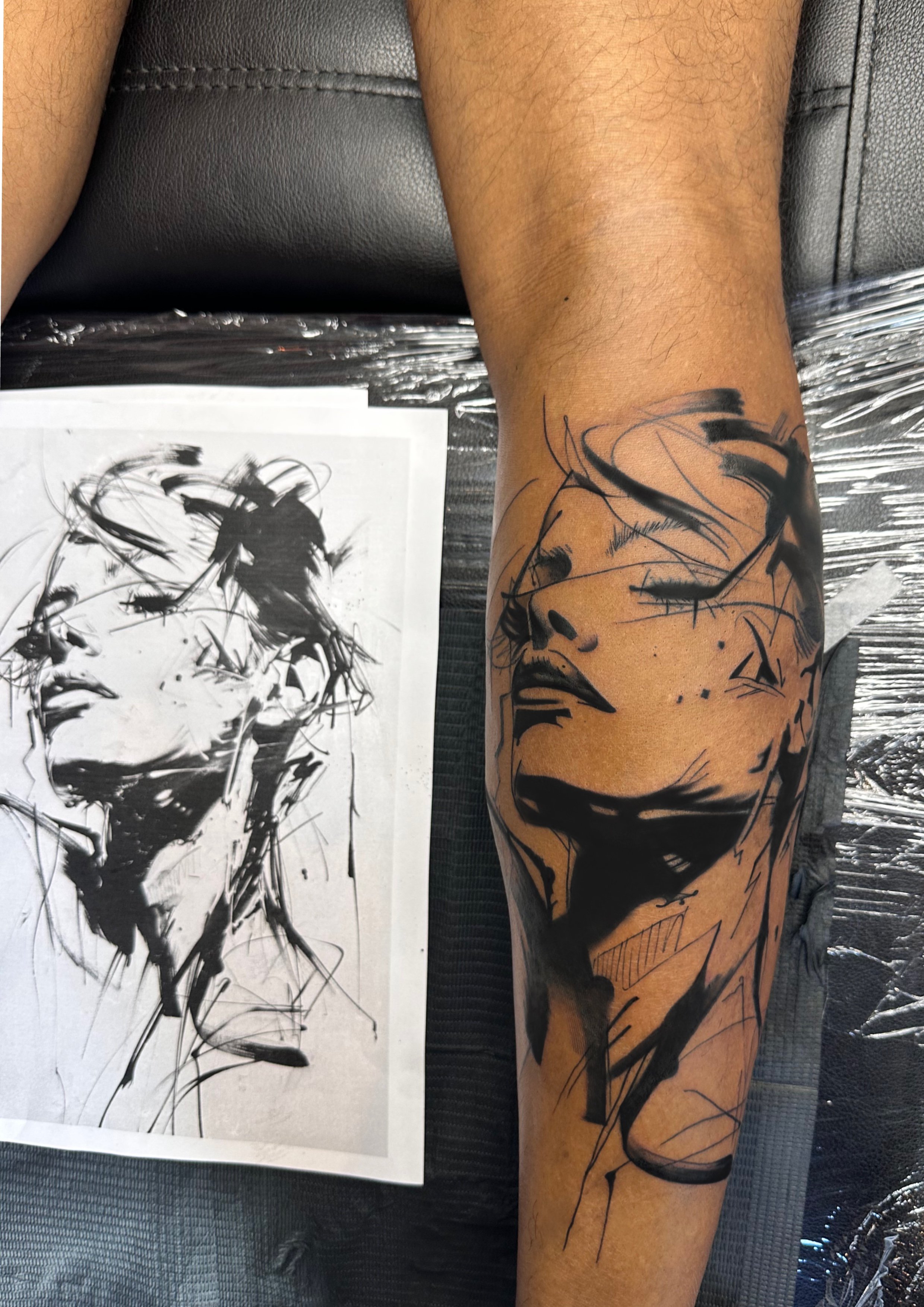 A person's leg with a black tattoo of a stylized portrait of a woman's face, shown in a sketchy, abstract style.