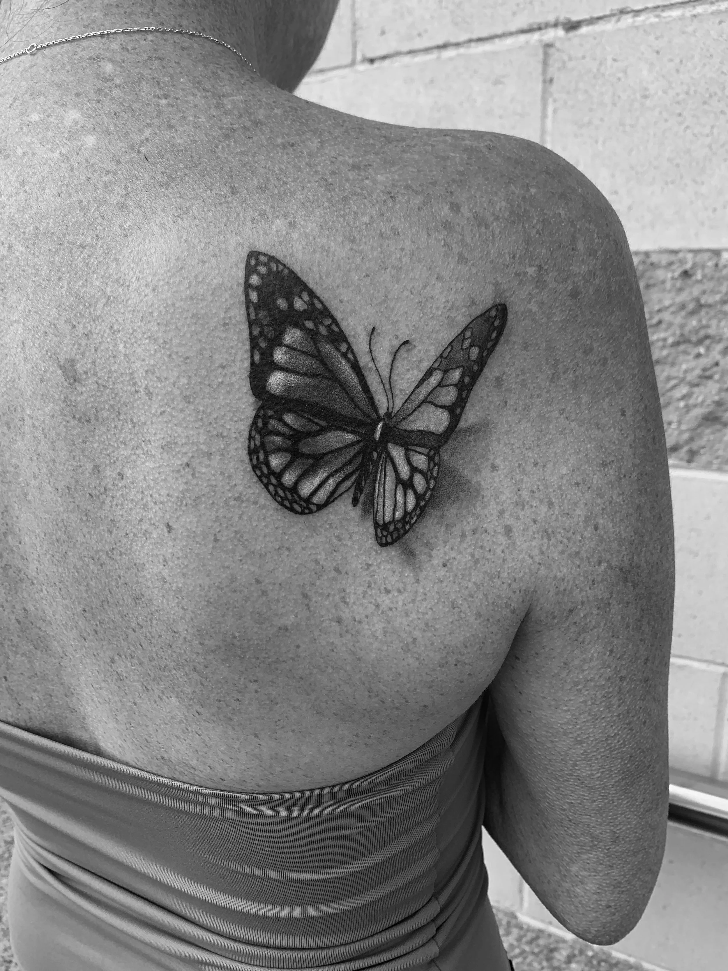 Black and white photograph of a person's upper back and shoulder, showing a butterfly tattoo on the shoulder blade.