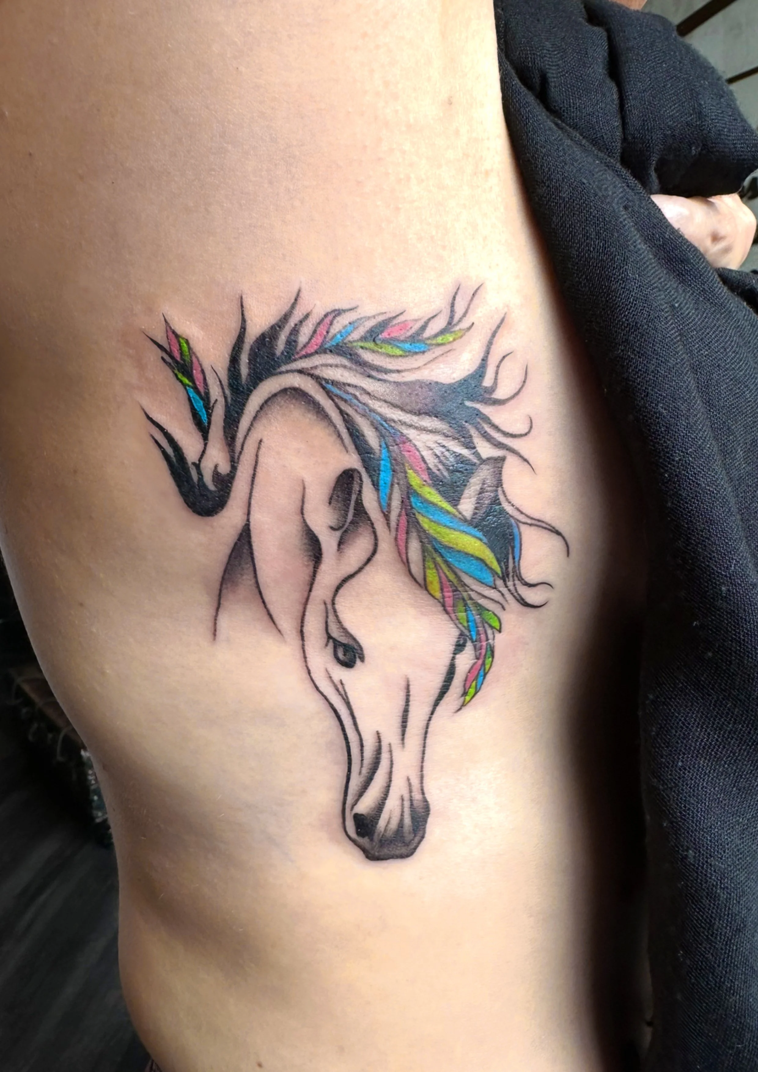 Tattoo of a stylized horse head with a flowing mane in black ink, decorated with colorful streaks of pink, blue, green, and yellow.