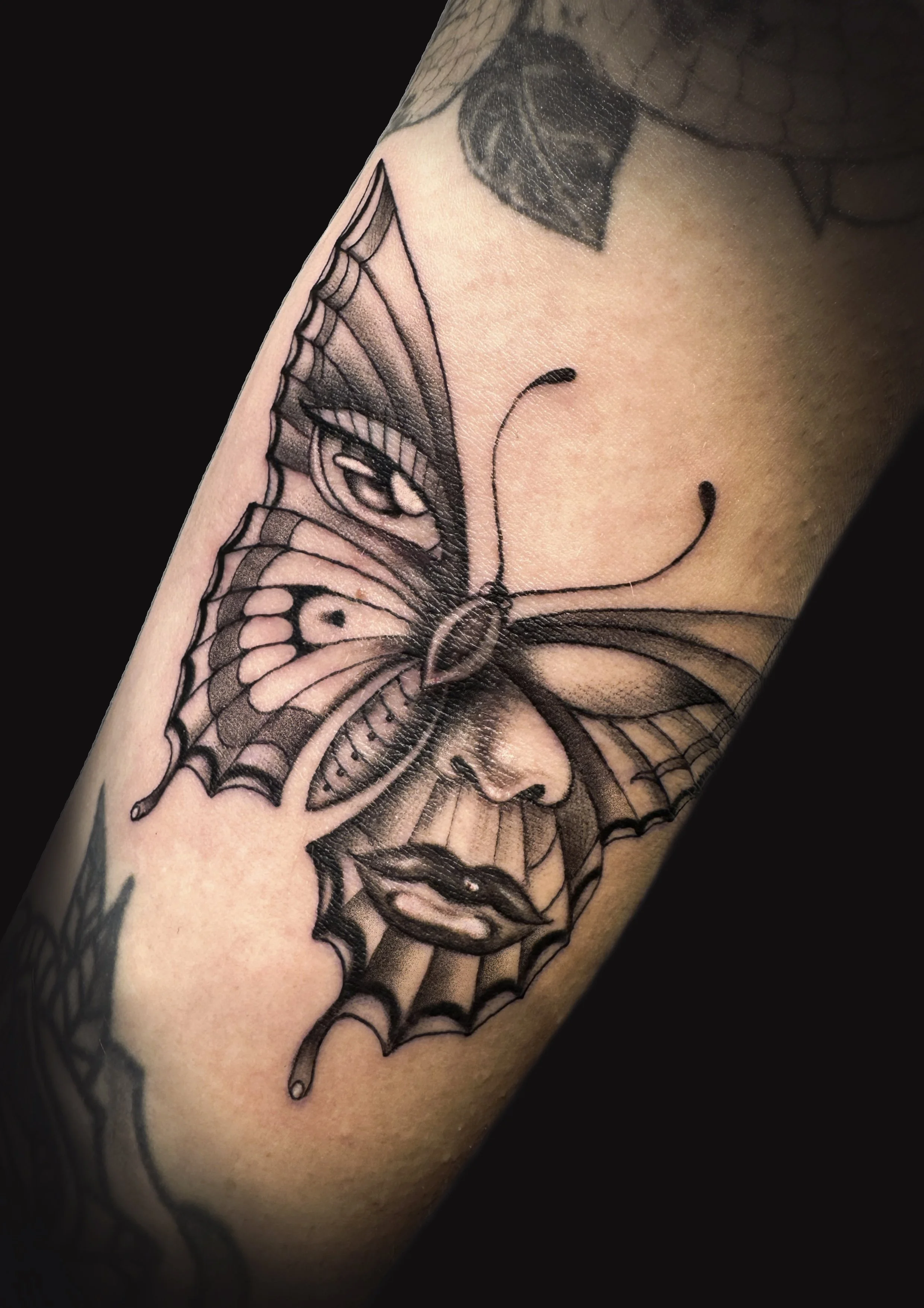 Black and gray tattoo of a butterfly with human facial features, including lips and nose, on skin.