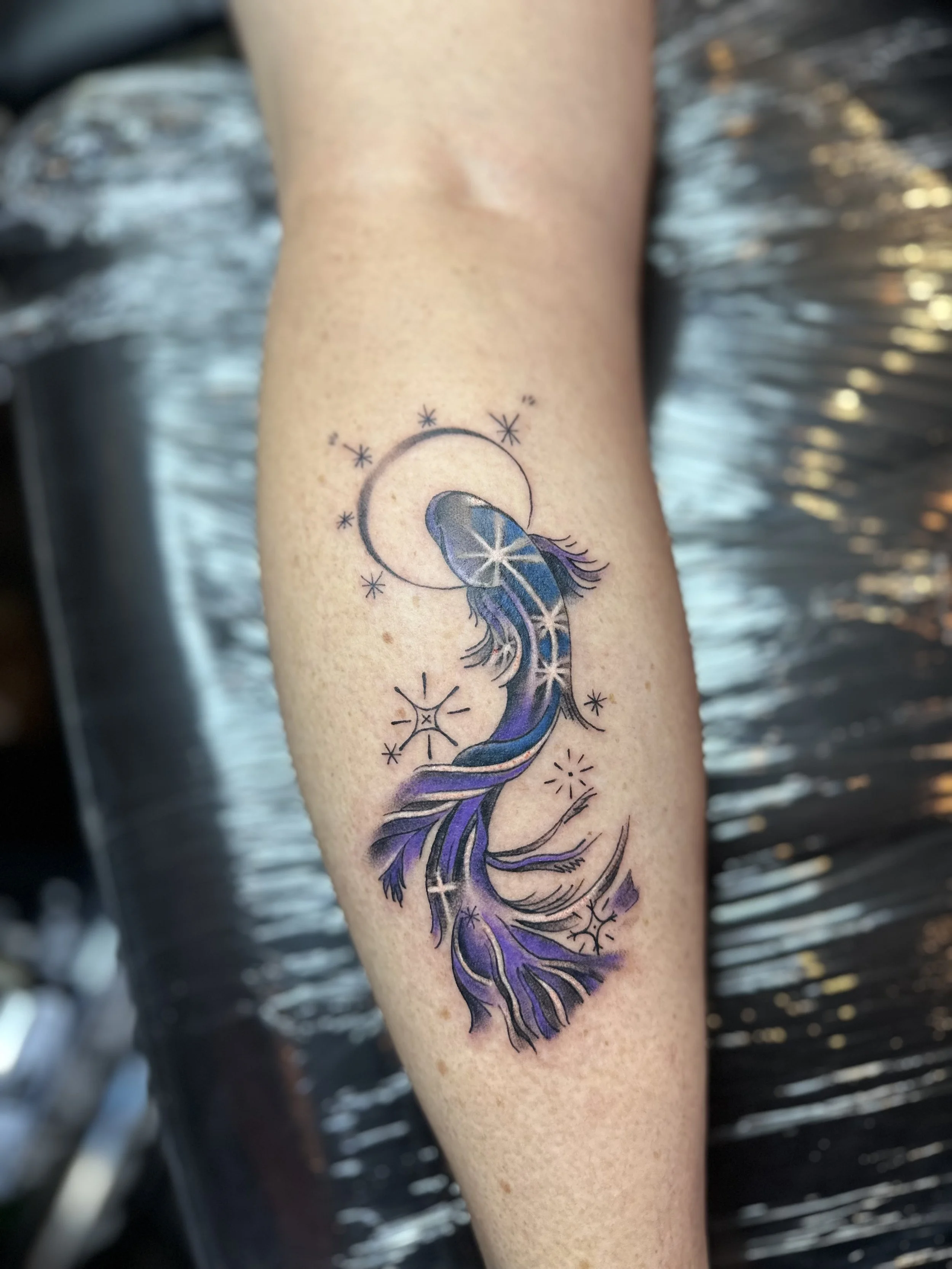 Tattoo of a purple and blue mermaid tail with star designs, moon, and celestial accents on a person's forearm.