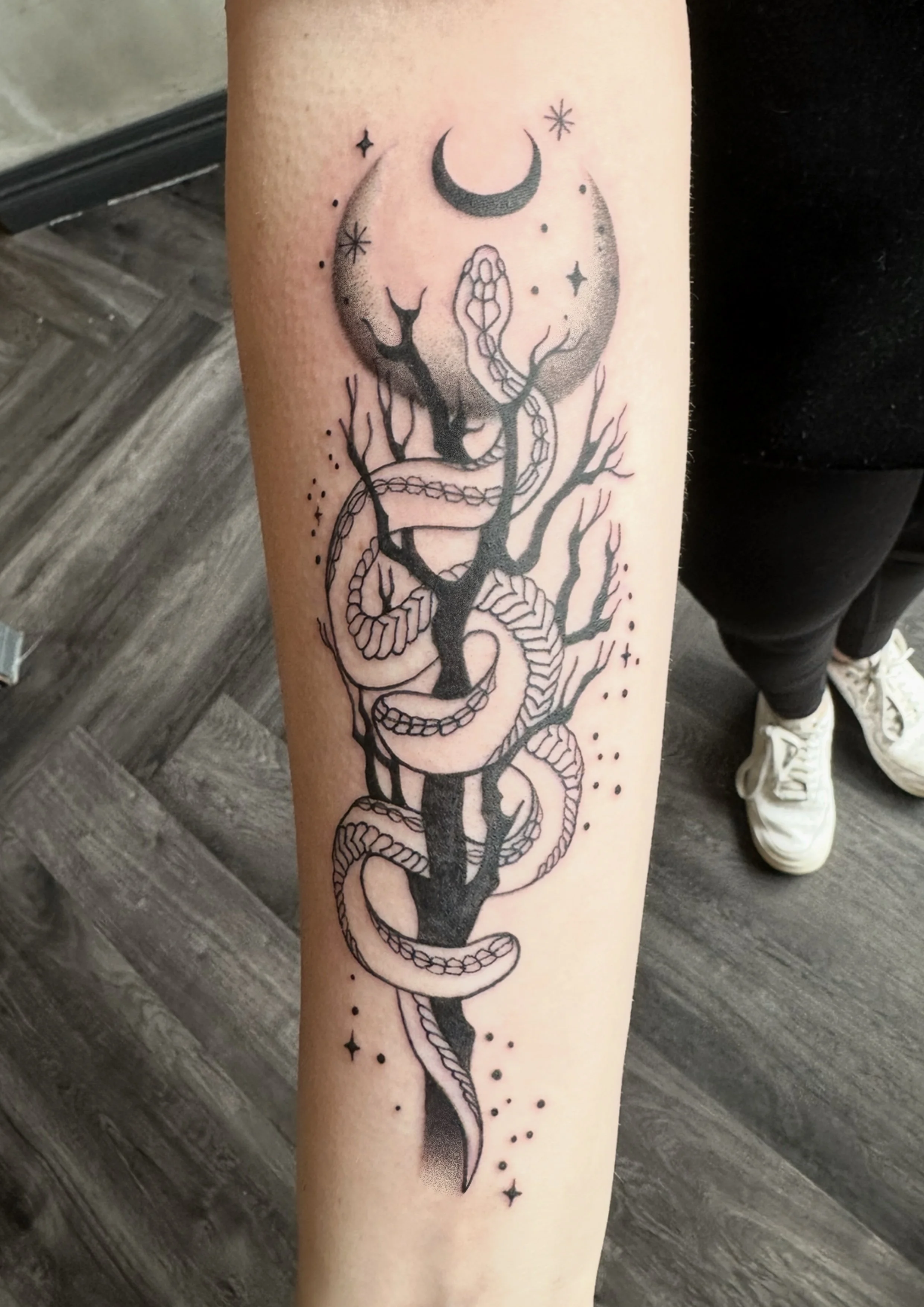 Tattoo of a black tree with bare branches, a snake coiled around the trunk, and a crescent moon above surrounded by stars and dots.