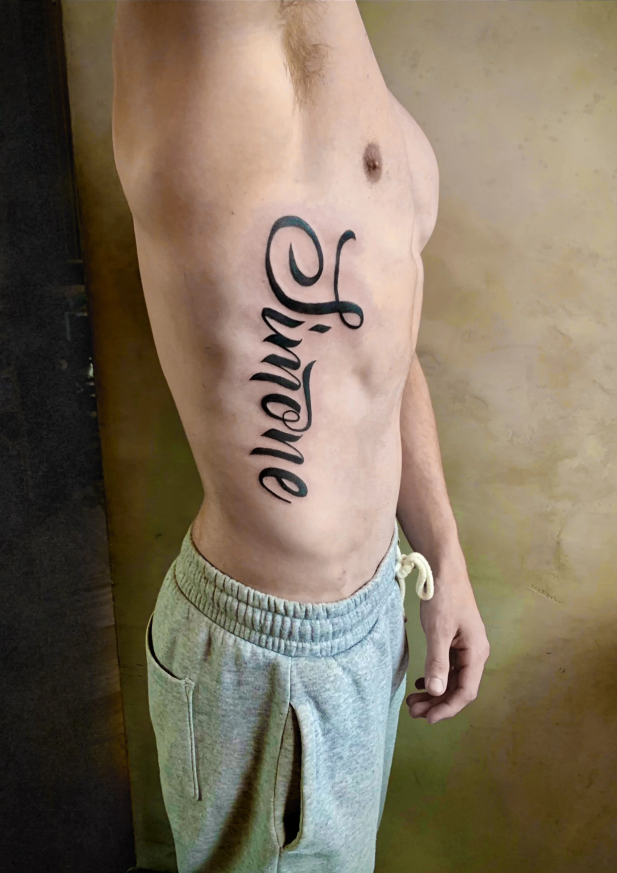 A shirtless man with a chest tattoo that reads 'Love' in cursive script, wearing light gray sweatpants.