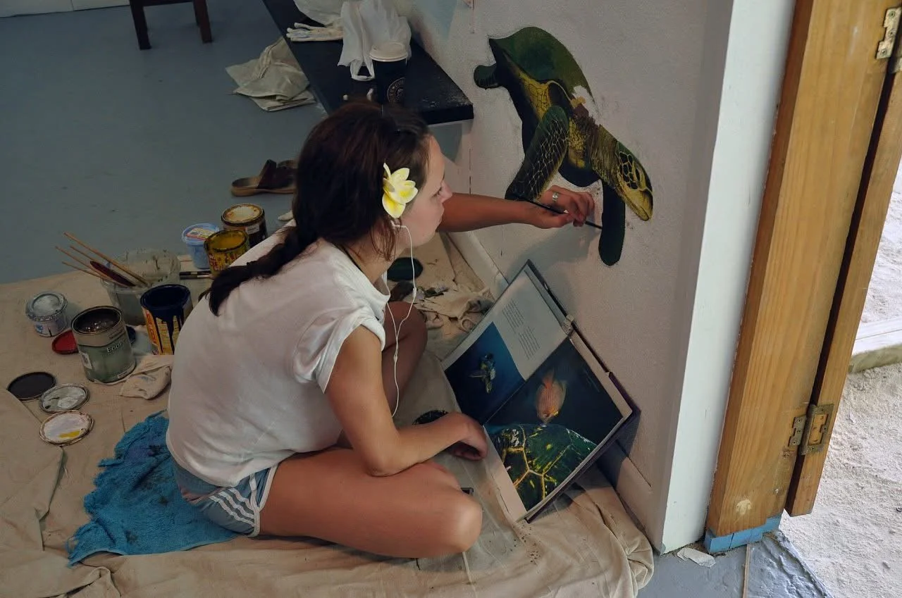 A woman painting a large, colorful lizard on a wall while sitting on the floor. She has a flower in her hair and there are art supplies around her, including open paint cans, brushes, and a book with underwater images.