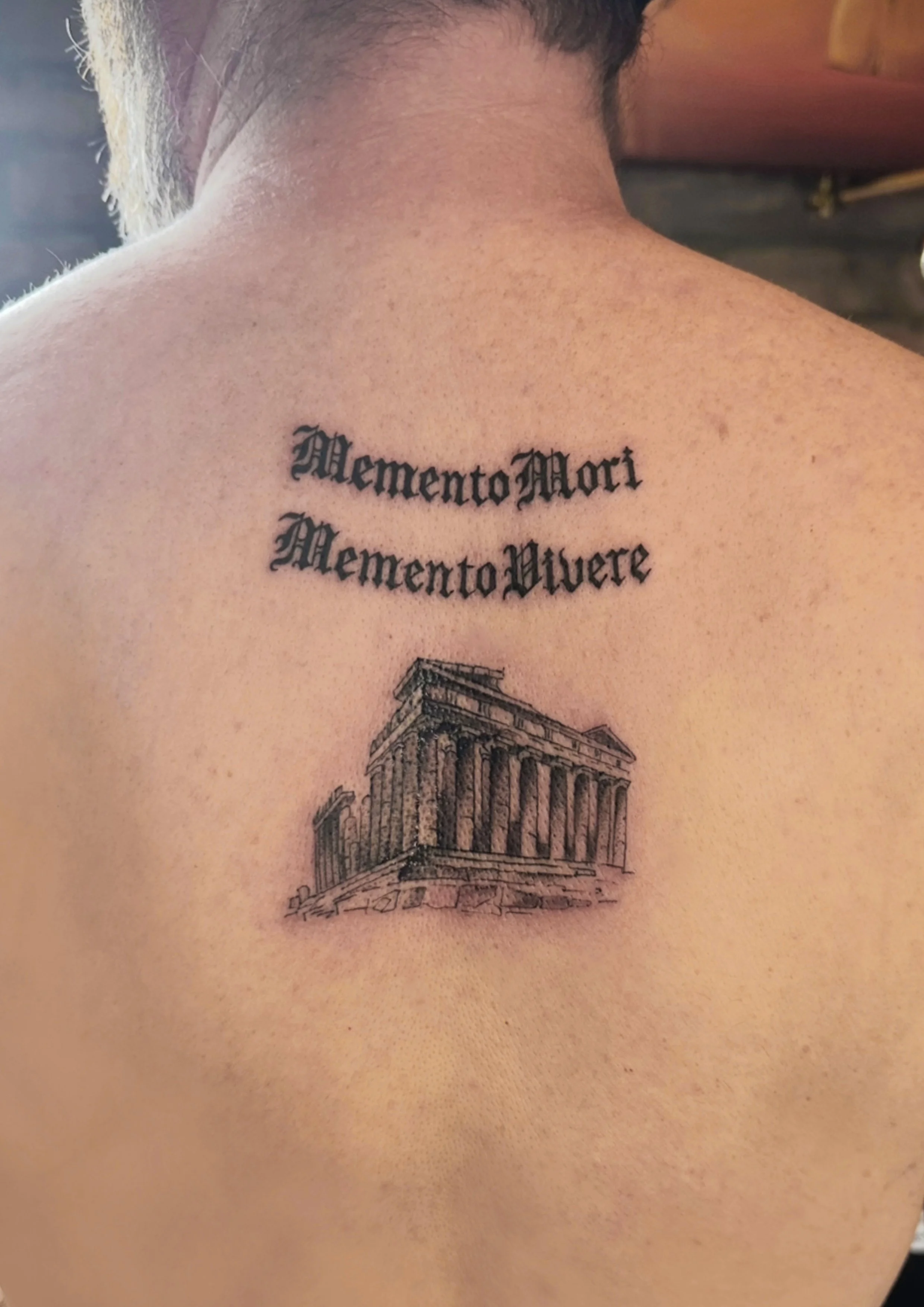 Tattoo on person's upper back with the phrase 'Memento Mori' written in Latin above an illustration of an ancient Greek temple.