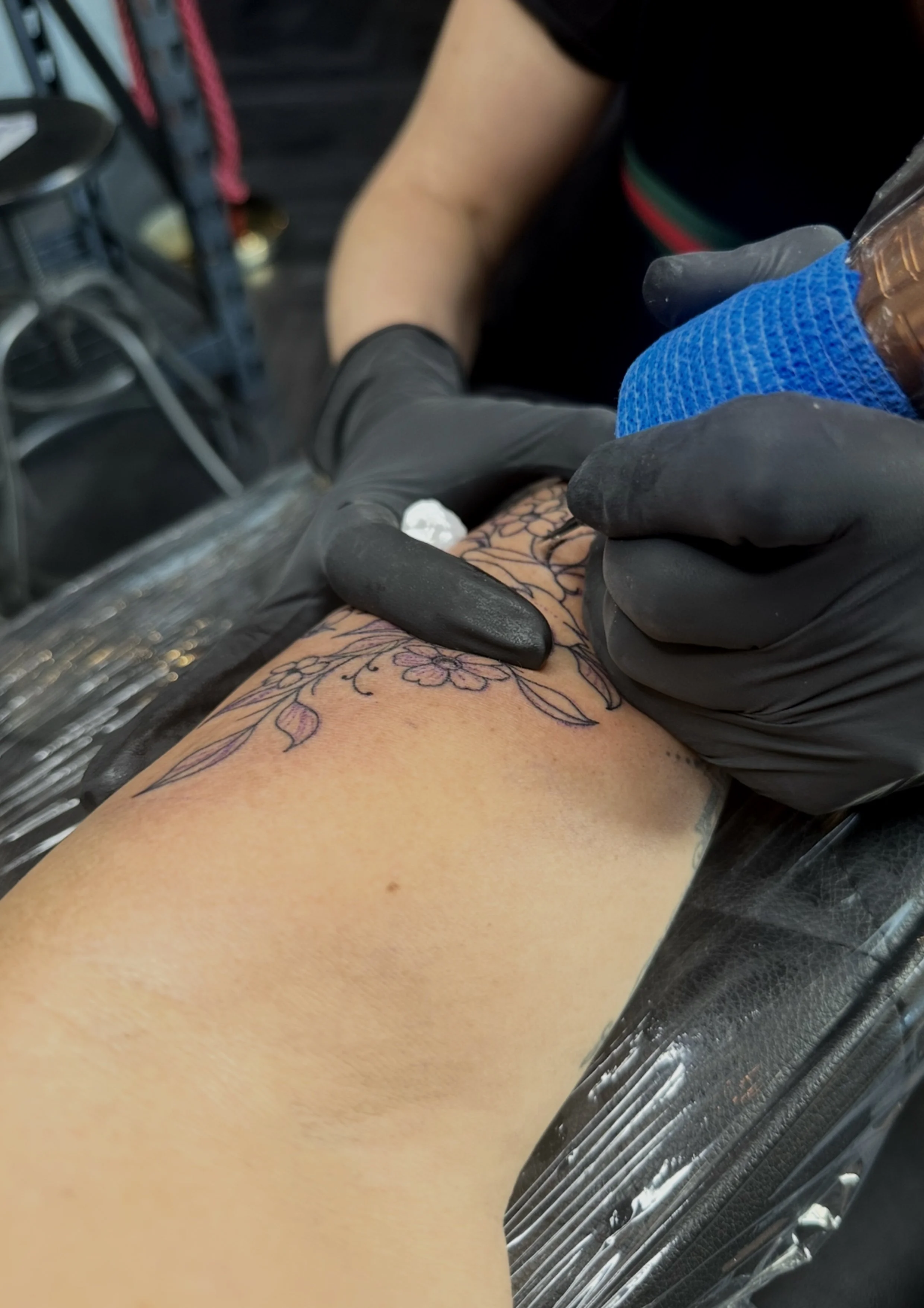 A person getting a tattoo on their forearm, with the tattoo artist using a tattoo machine. The artist is wearing black gloves and the tattoo design features flowers and leaves.