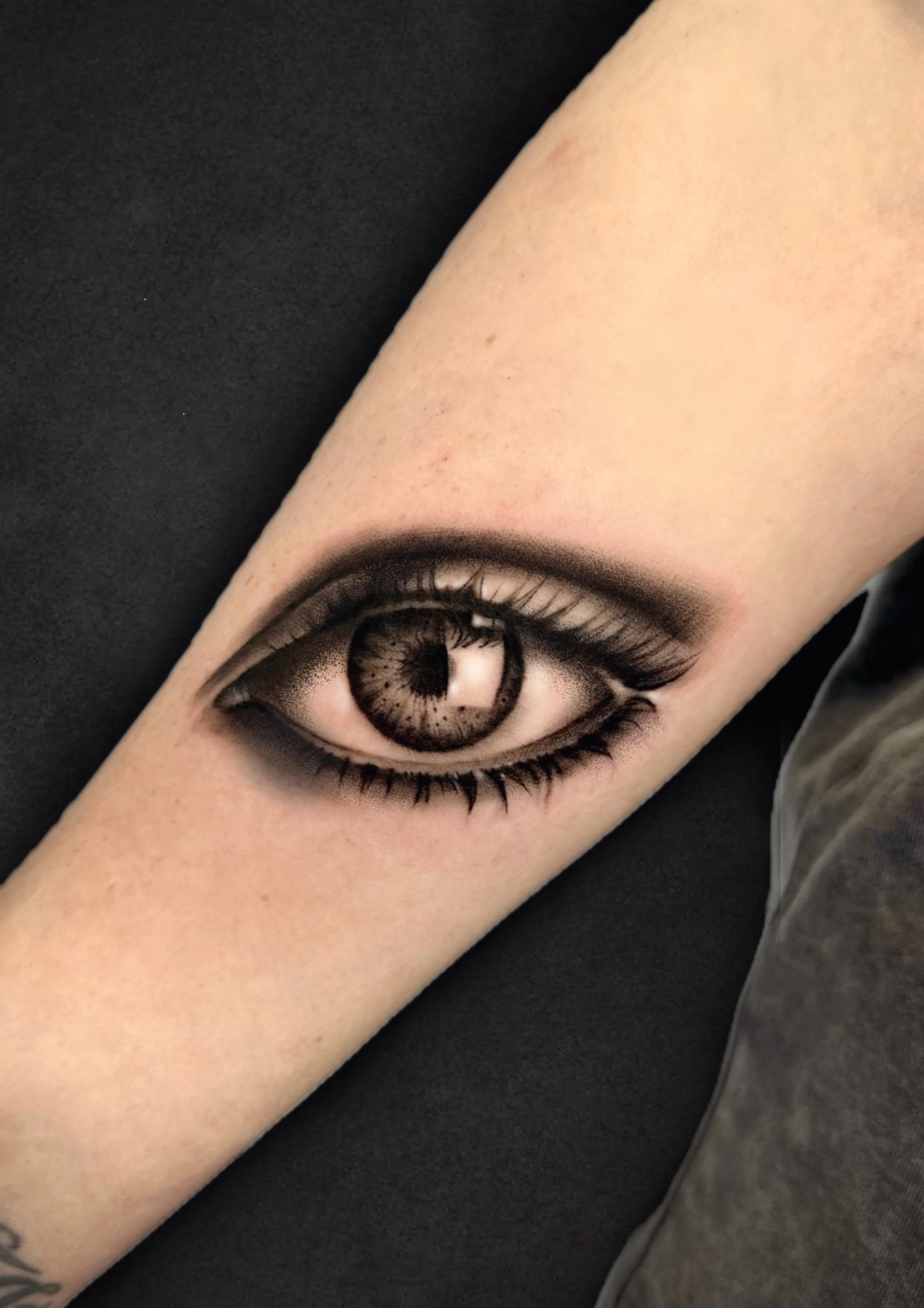 A tattoo of a realistic eye on an inner forearm, with detailed eyelashes and eyebrow, against a black background.