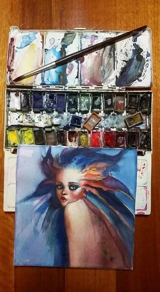 A watercolor painting of a woman's face with colorful, flowing hair set against a blue background, surrounded by a watercolor palette with various colors and a paintbrush.