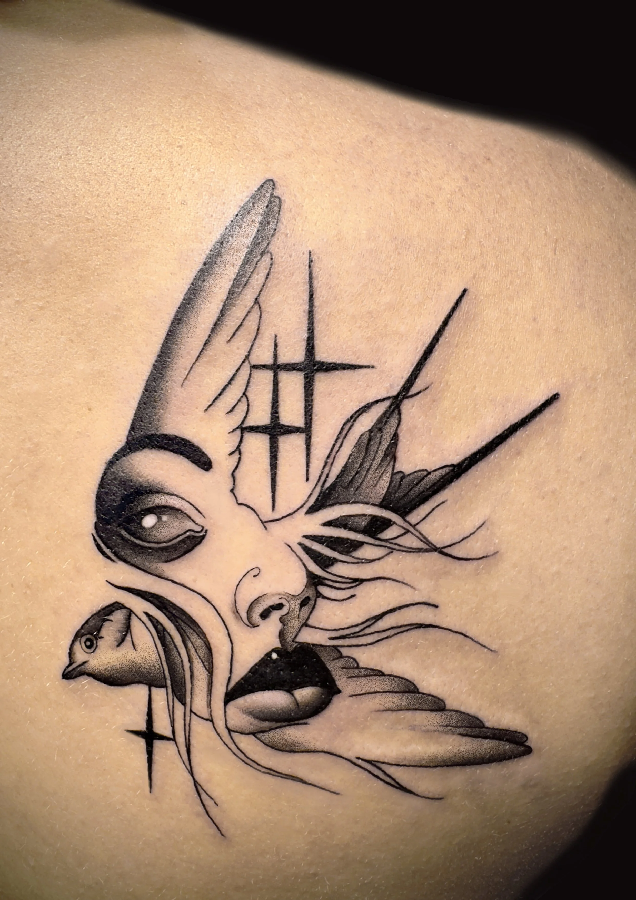 Black and gray tattoo of a woman's face with bird features and abstract lines, stars, and geometric shapes.
