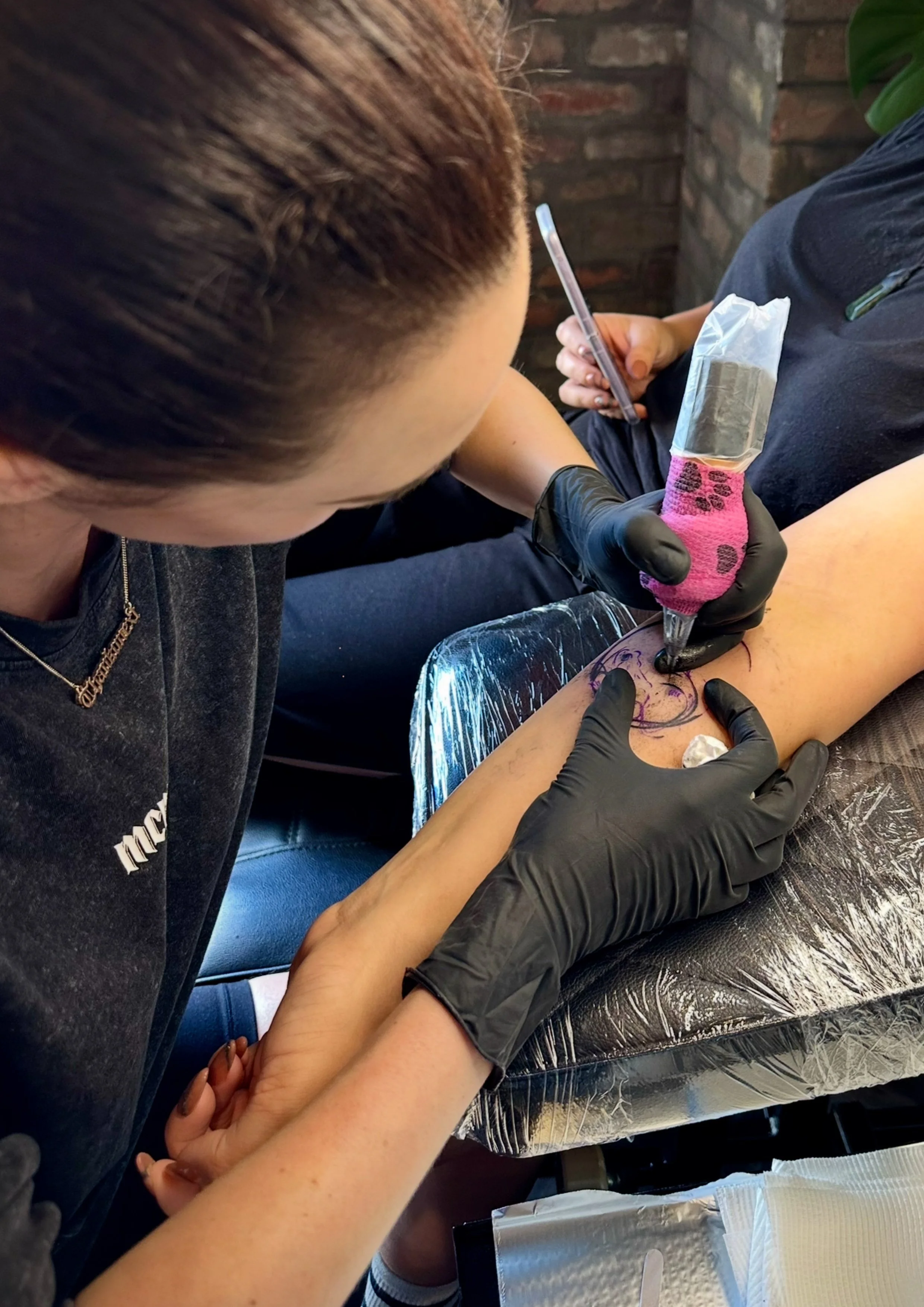 A tattoo artist is creating a design on a person's forearm using ink and a tattoo machine. The artist is wearing gloves and is focused on the work, with a phone in the other hand.