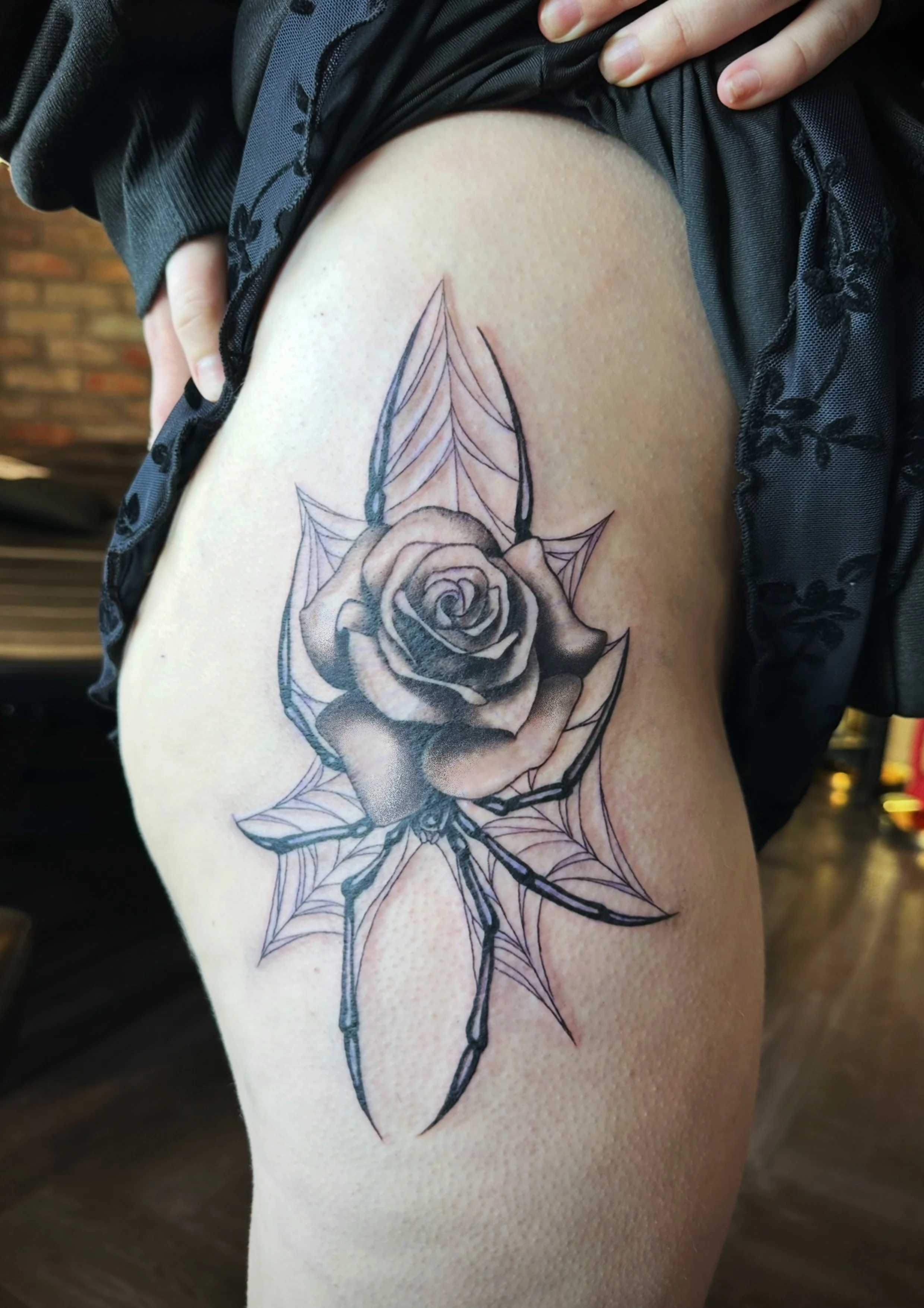 A black and gray tattoo of a rose with spider web details on the leaves, located on a person's thigh.