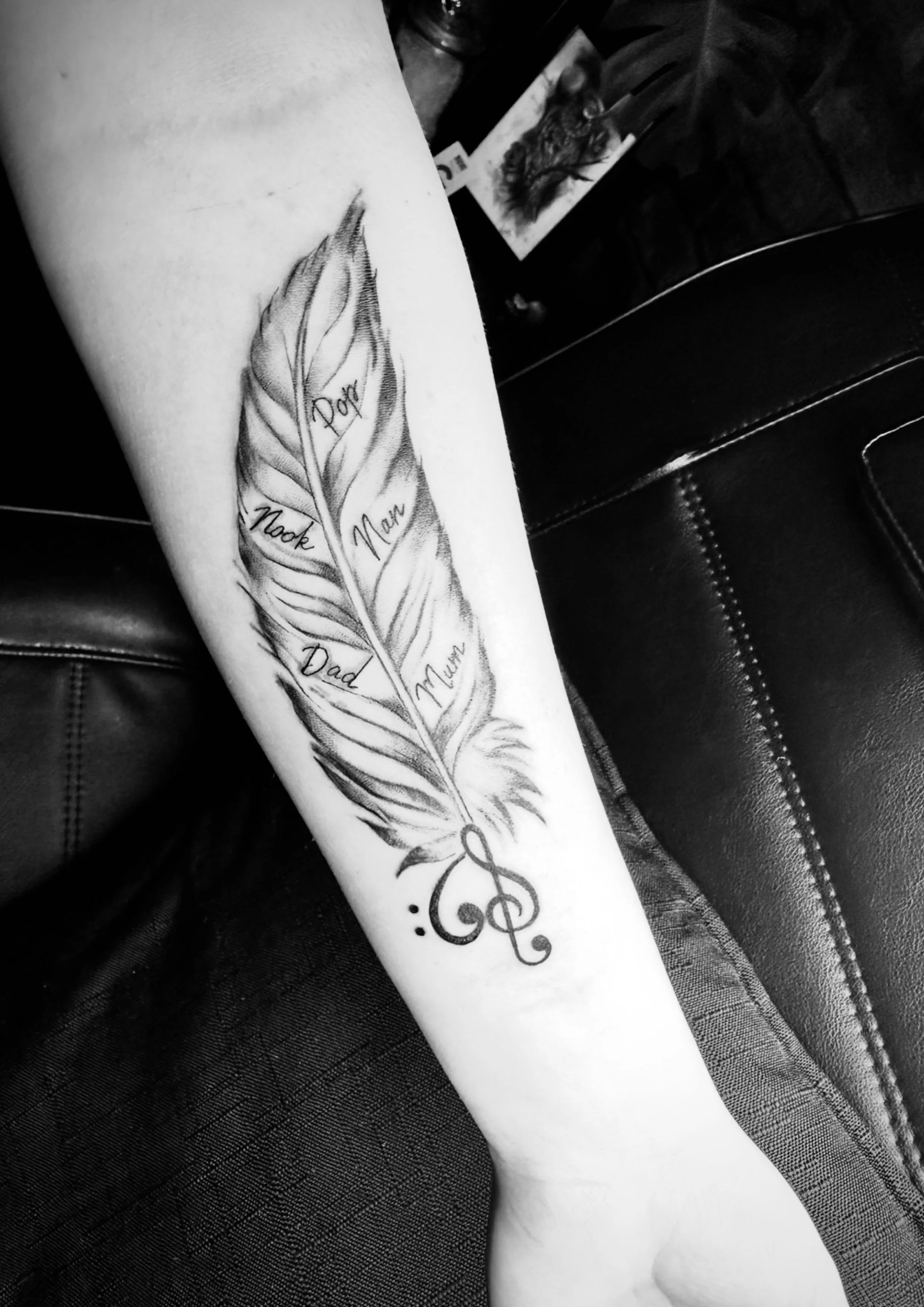 Tattoo of a feather with written names of family members and a treble clef at the bottom.