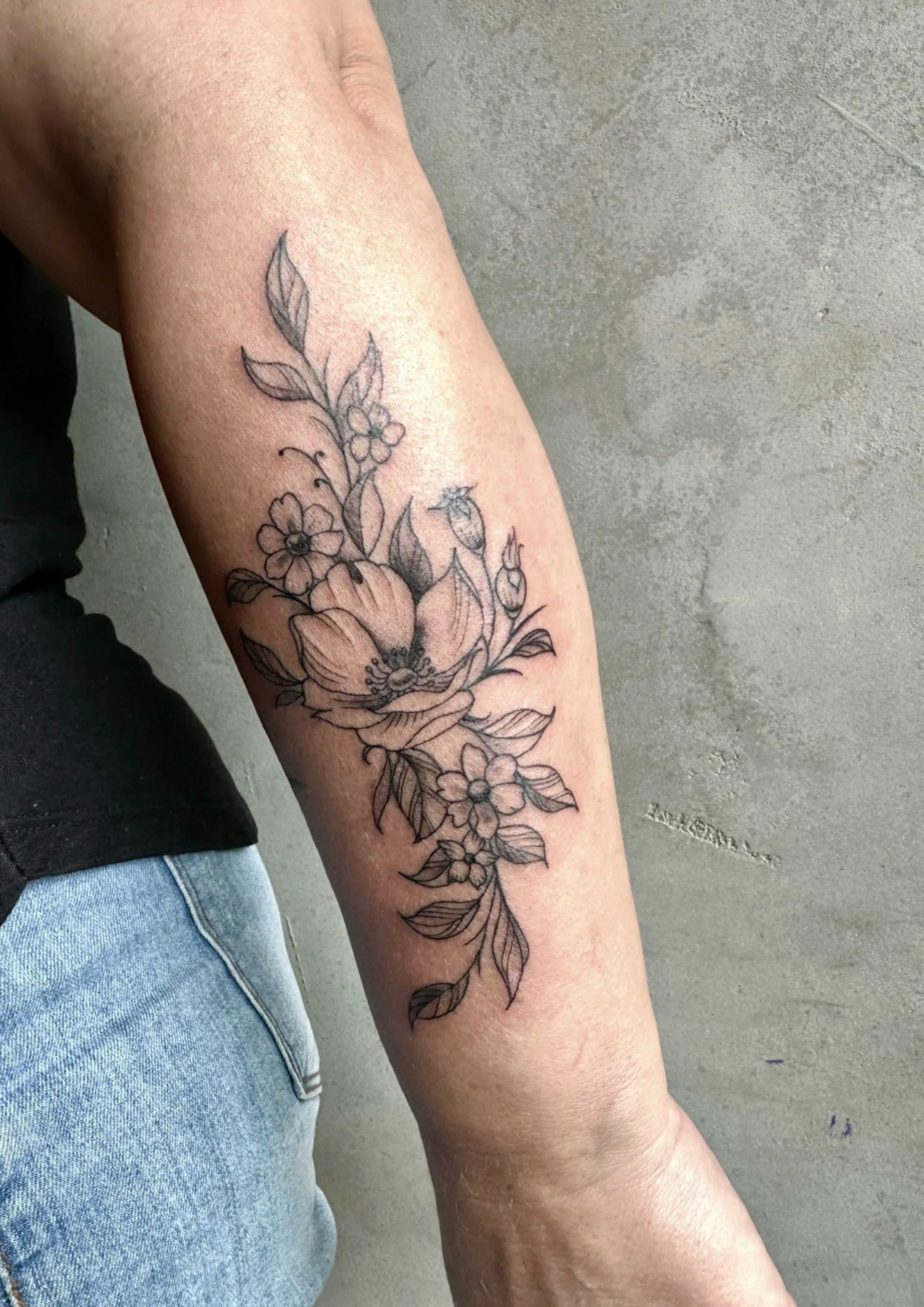 Black outline floral tattoo on the forearm, featuring large flowers, smaller blossoms, and leafy branches.