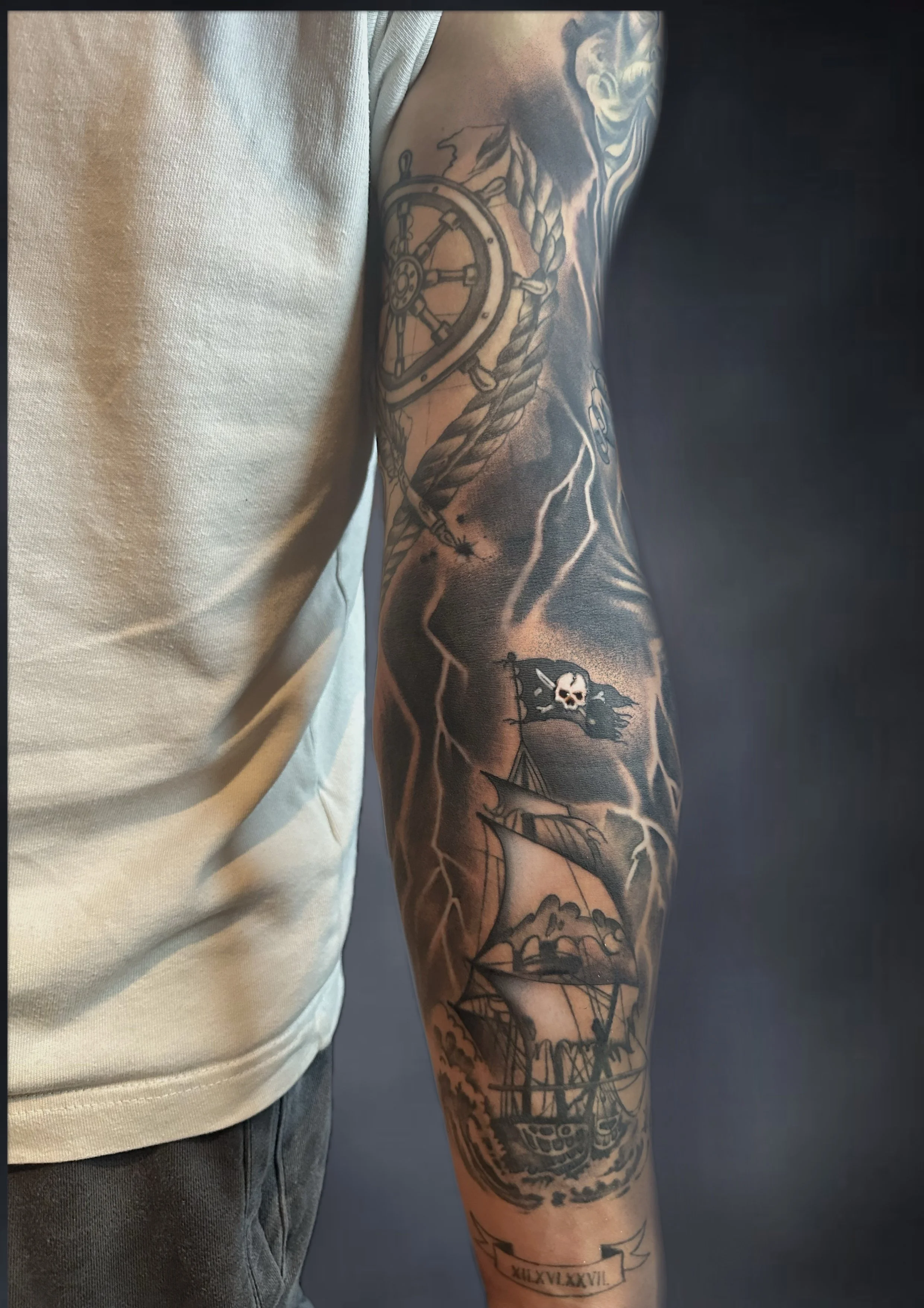 Black and gray tattoo sleeve featuring a ship with sails, a pirate flag with a skull, a compass, and lightning bolts on the arm.